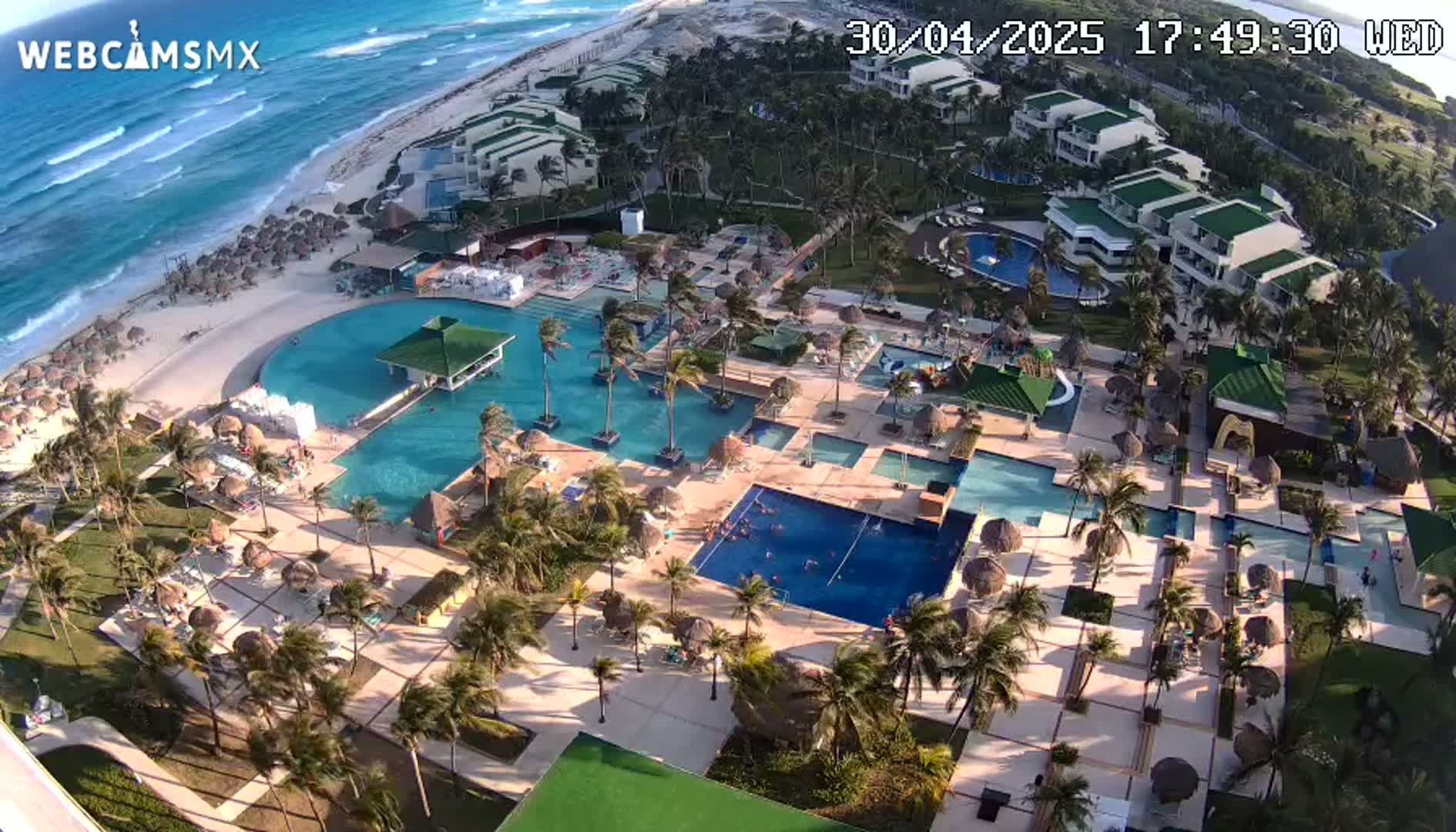 An aerial view shows a resort with numerous pools and buildings situated next to a beach with wavy ocean under a sunny sky.