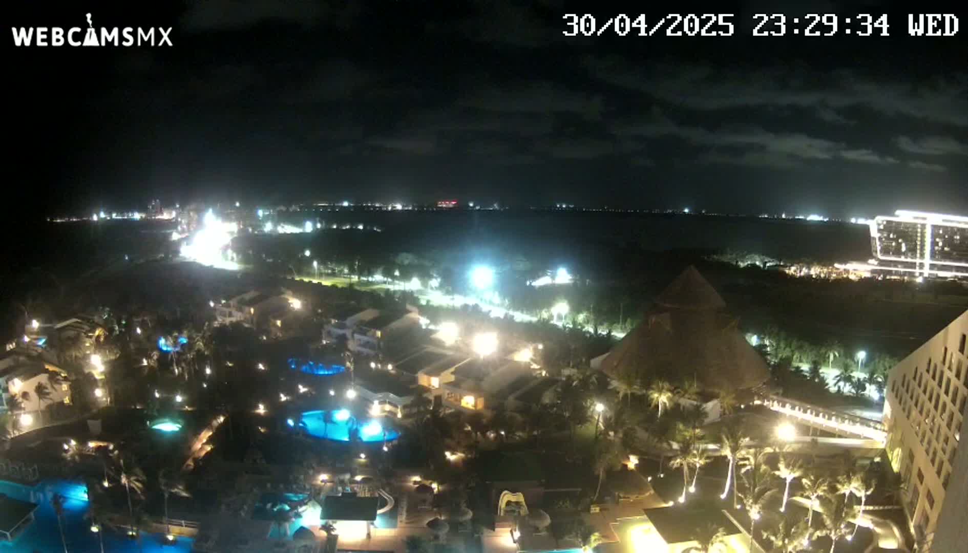 A nighttime view of a resort complex with illuminated pools and buildings, palm trees, and a body of water in the distance under a partly cloudy sky.