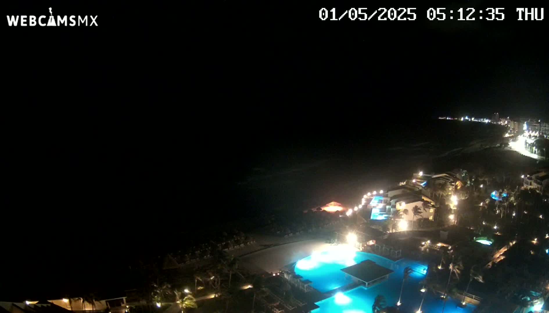 A nighttime aerial view shows a coastal resort with illuminated pools and buildings, overlooking a dark ocean under a clear, starless sky.