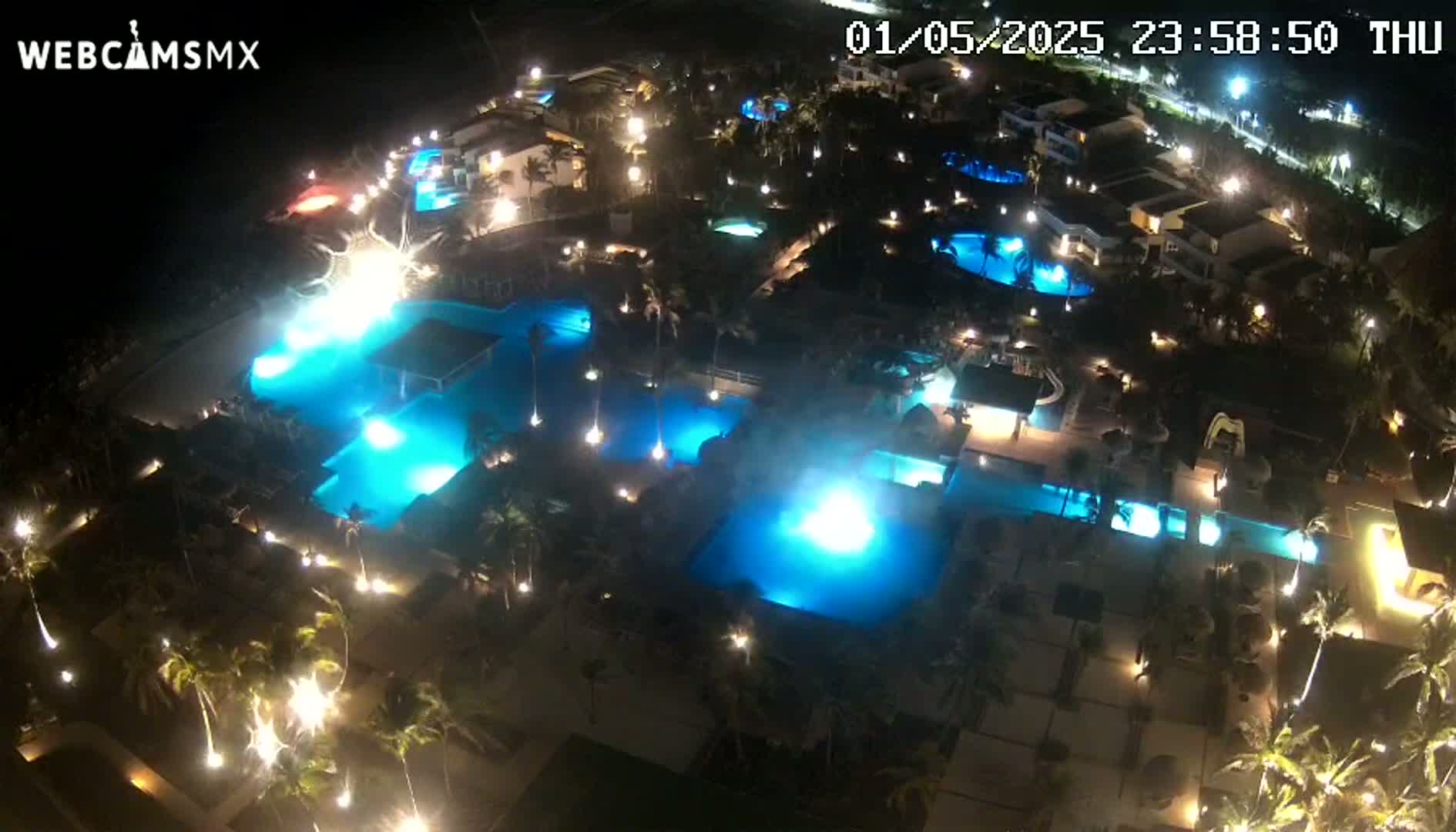 An aerial nighttime view shows numerous illuminated swimming pools surrounded by palm trees and buildings.