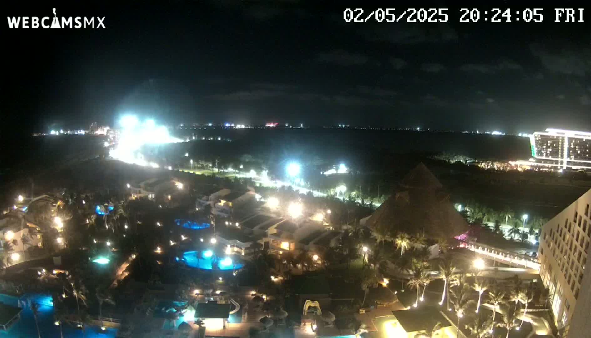 A nighttime view of a resort area with illuminated buildings, pools, and landscaping, overlooking a dark ocean under a clear sky.