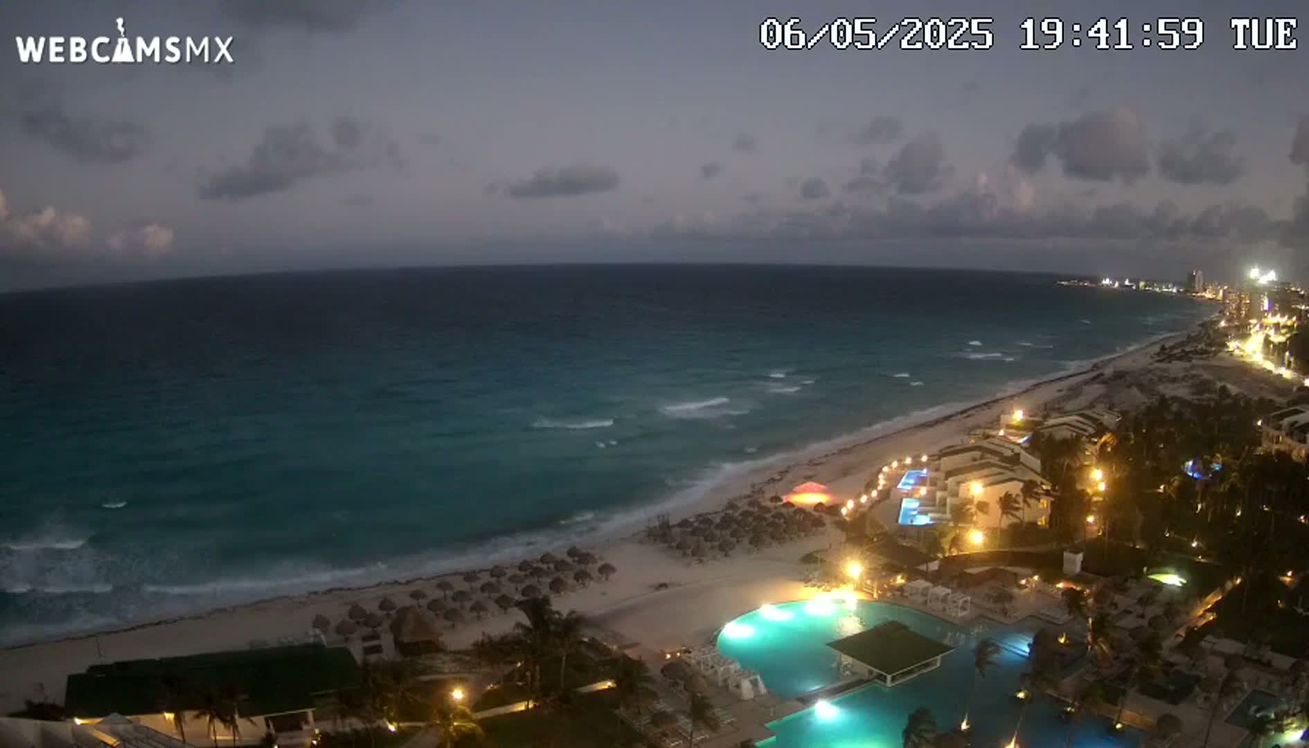 A nighttime view of a beach with gently crashing waves, illuminated buildings, and swimming pools under a partly cloudy sky.