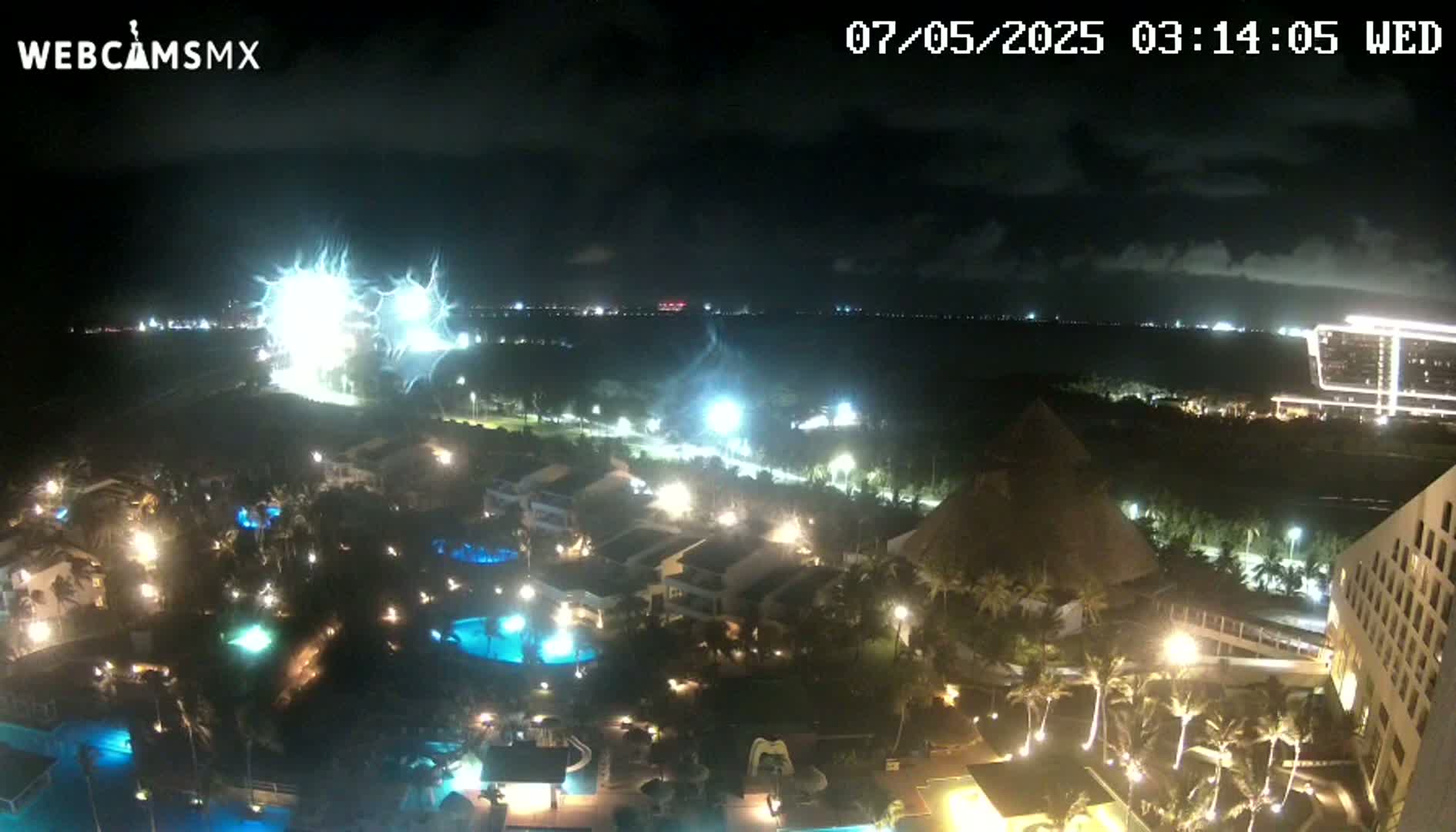 A nighttime, aerial view of a resort complex with illuminated pools and buildings, under a partly cloudy sky, with bright lights possibly from a fireworks display visible in the distance.