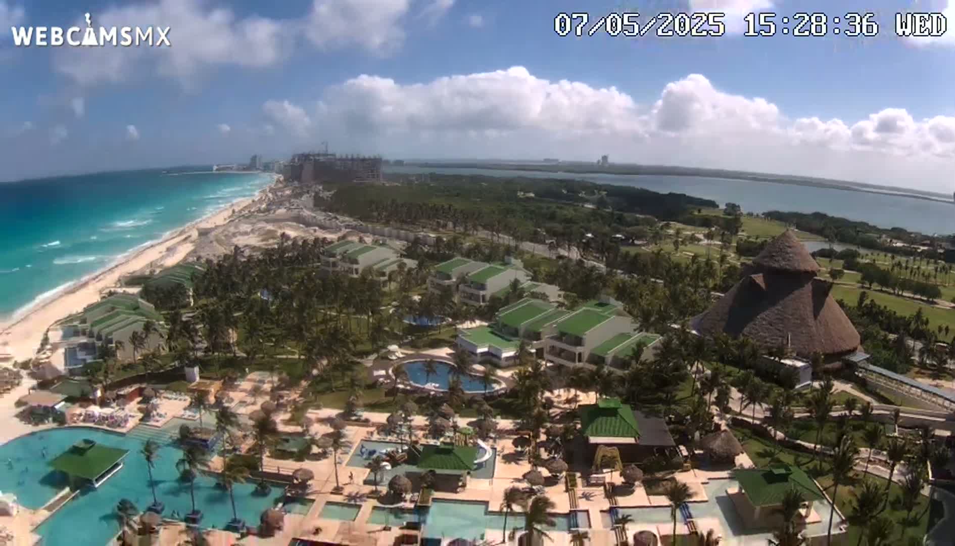 An aerial view shows a resort with multiple pools and buildings on a beach, under partly cloudy skies, with a body of water and more distant buildings visible in the background.