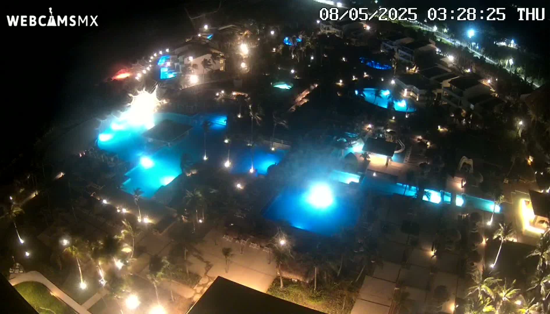 An aerial nighttime view shows numerous illuminated swimming pools surrounded by palm trees and buildings.