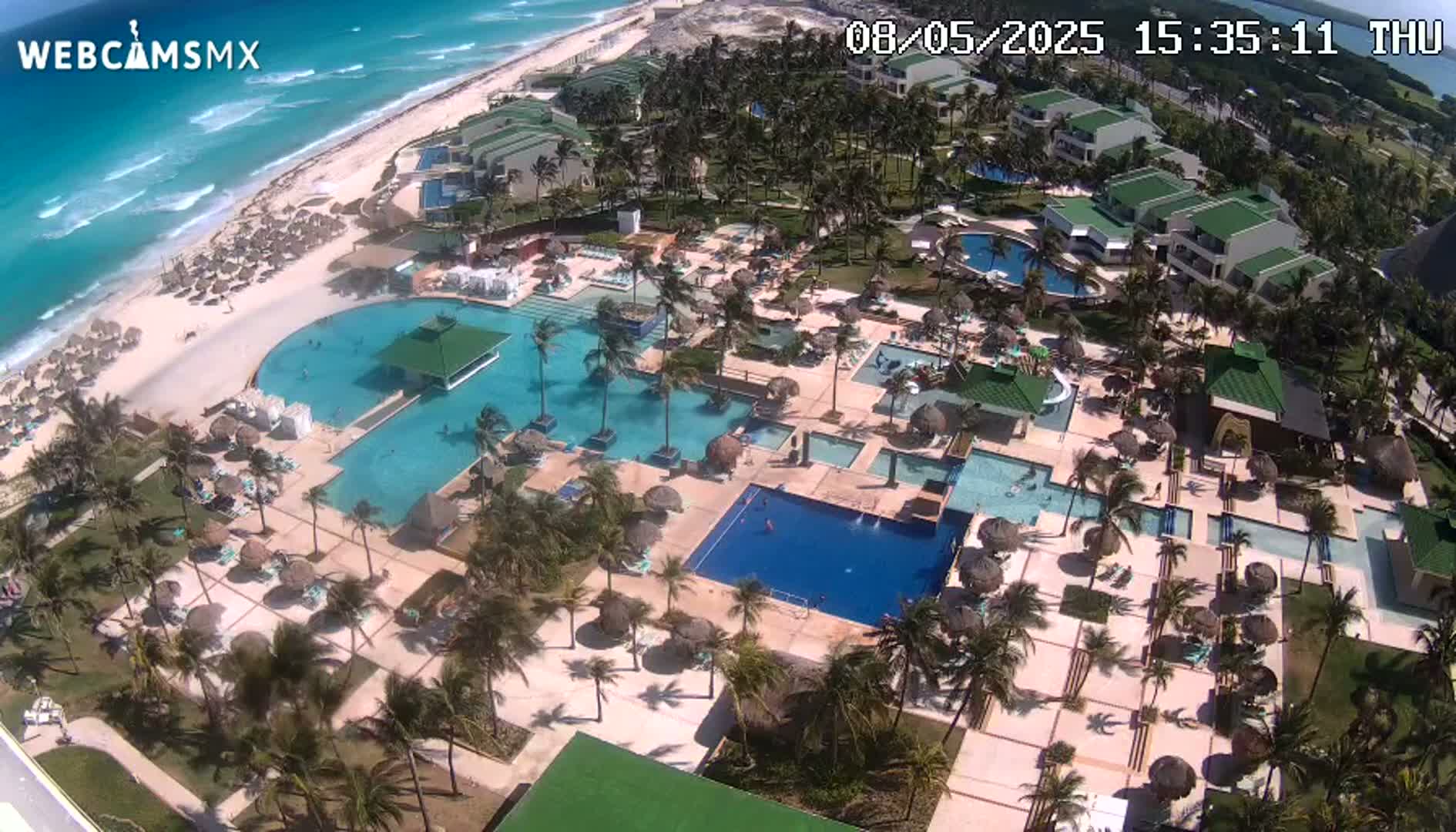 An aerial view shows a resort with numerous pools, palm trees, and beachside buildings on a sunny day with a bright blue ocean in the background.