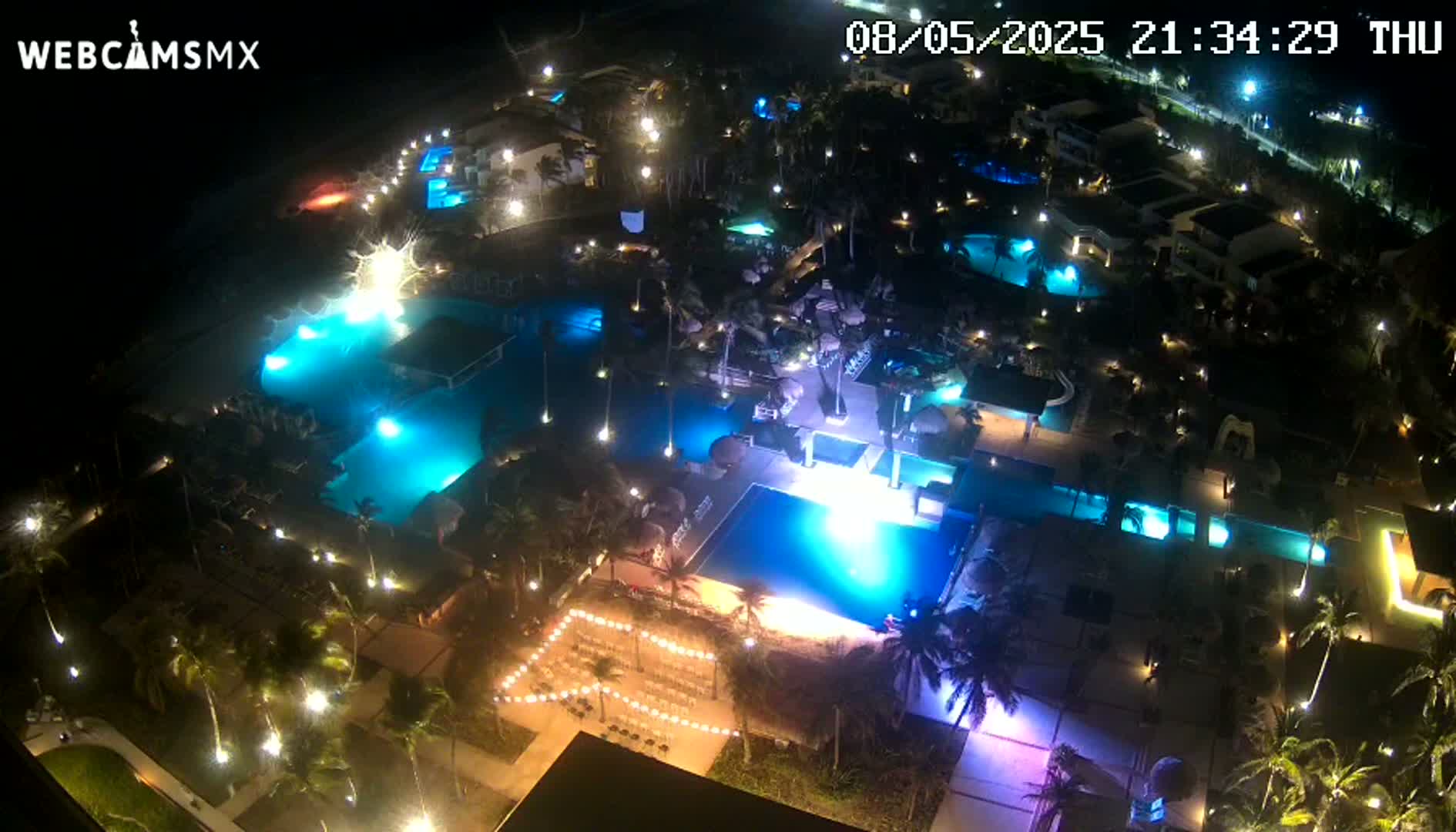An aerial nighttime view shows a resort with numerous illuminated pools and palm trees surrounding a wedding setup.