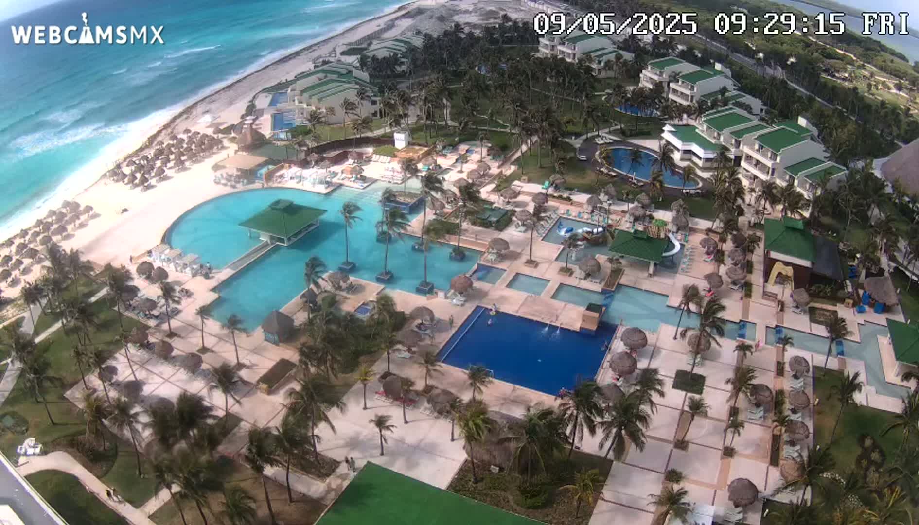An aerial view of a resort with multiple pools, palm trees, and a beach with turquoise water under partly cloudy skies.
