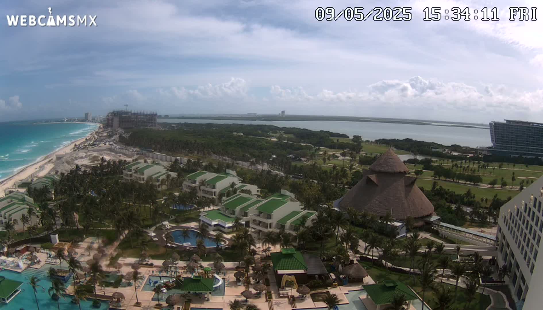 A partly cloudy day shows an aerial view of a tropical resort with pools, buildings, and lush landscaping next to a beach and ocean, with additional resorts and construction visible in the distance.