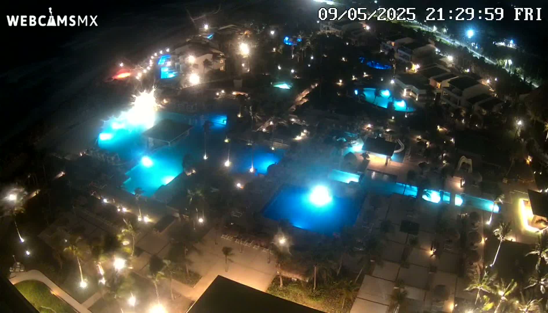 An aerial nighttime view shows a resort complex with numerous illuminated swimming pools and surrounding buildings.