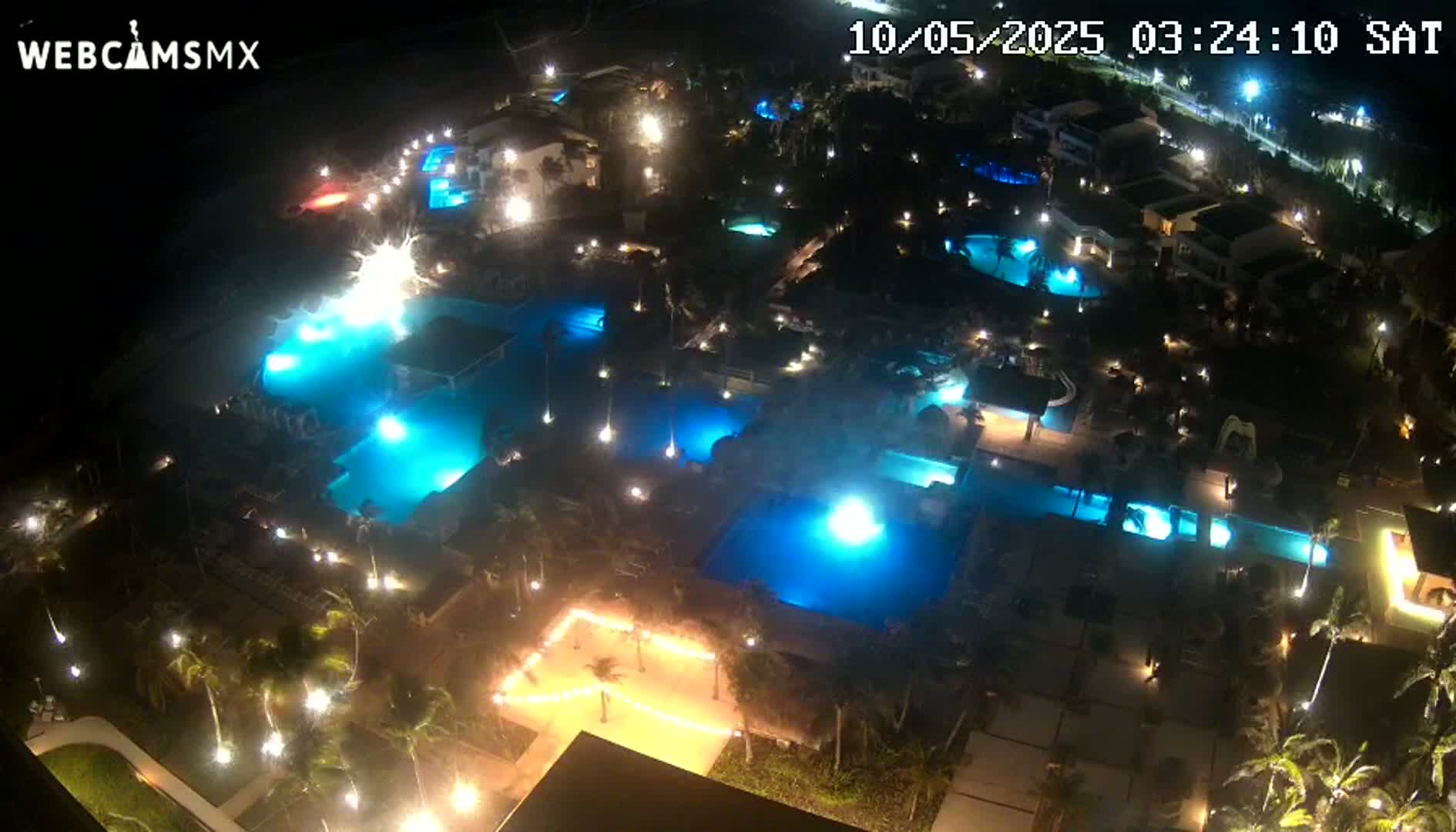 A nighttime aerial view shows numerous illuminated swimming pools and buildings surrounded by palm trees.