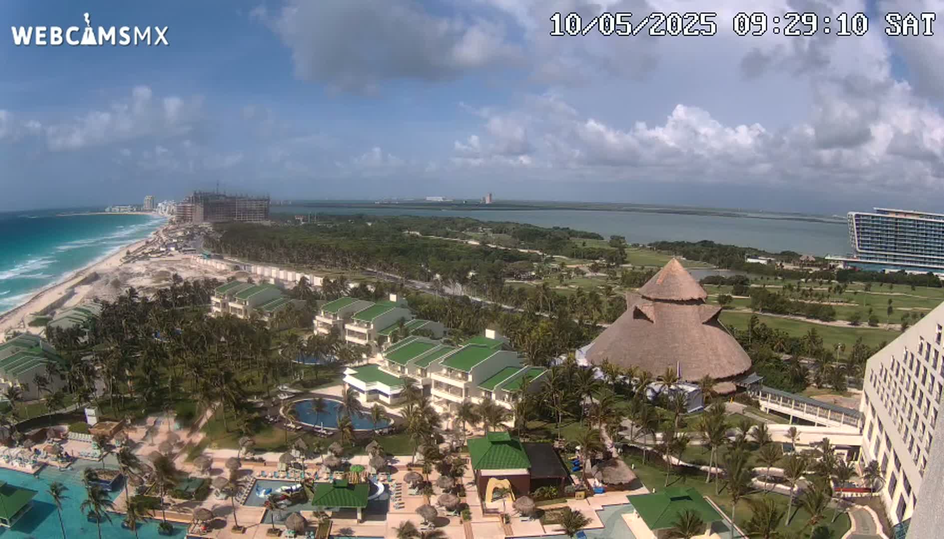 An aerial view shows a resort complex with pools and palm trees next to a beach and ocean under a partly cloudy sky.