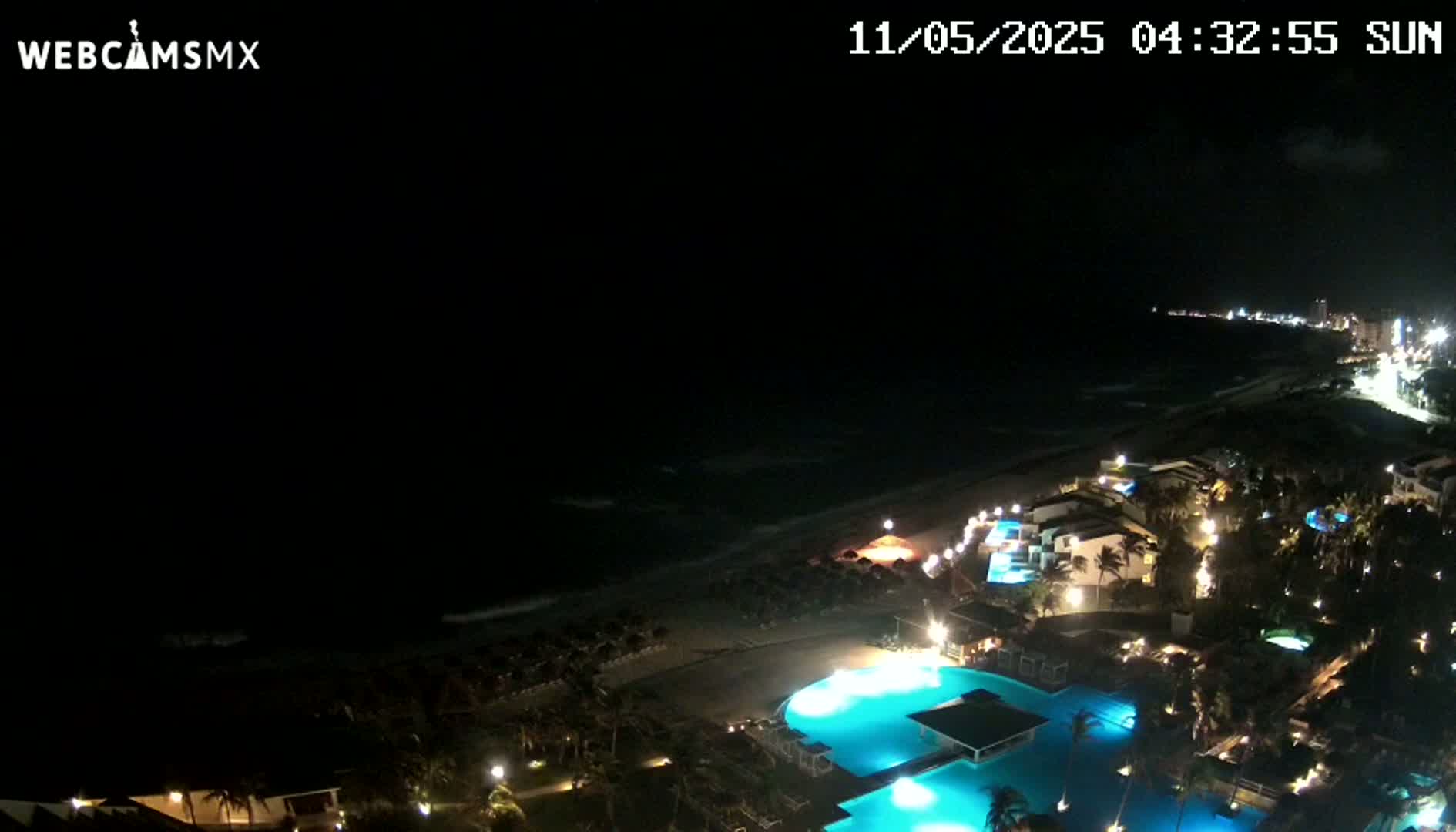 A nighttime view of a resort area with illuminated pools and buildings along a dark ocean shore under a clear sky.