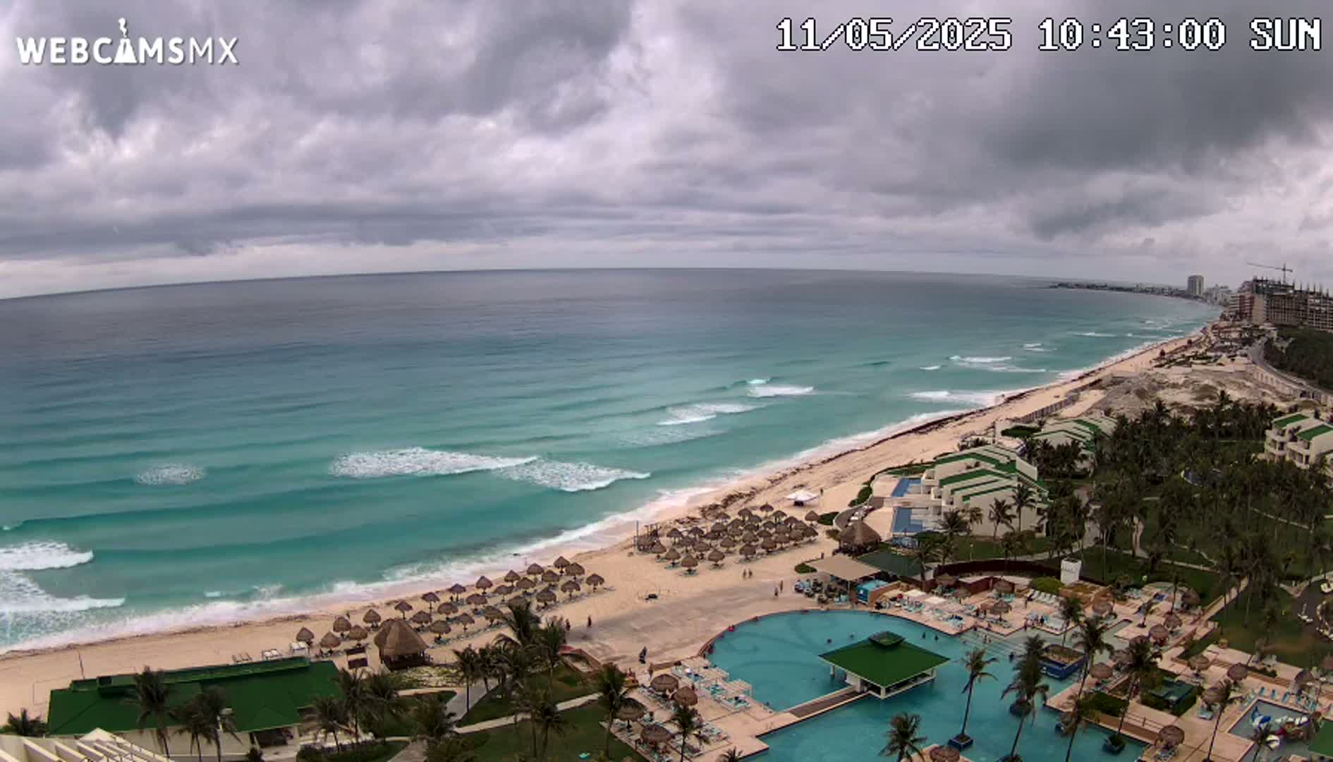 An overcast sky hangs over a beach resort with a turquoise ocean featuring gentle waves.