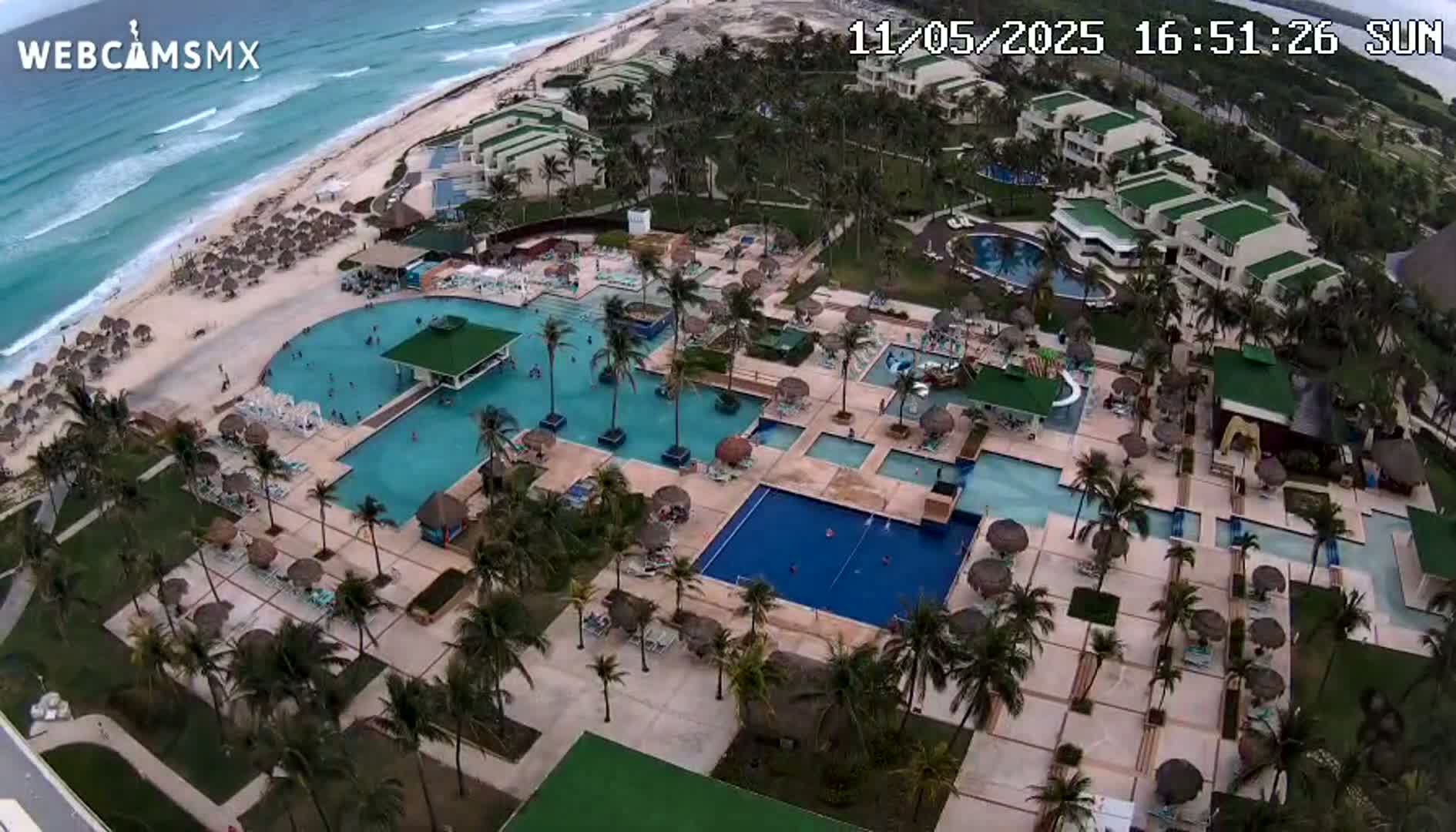 An aerial view shows a resort with numerous pools and palm trees, situated next to a beach with light waves under a partly sunny sky.