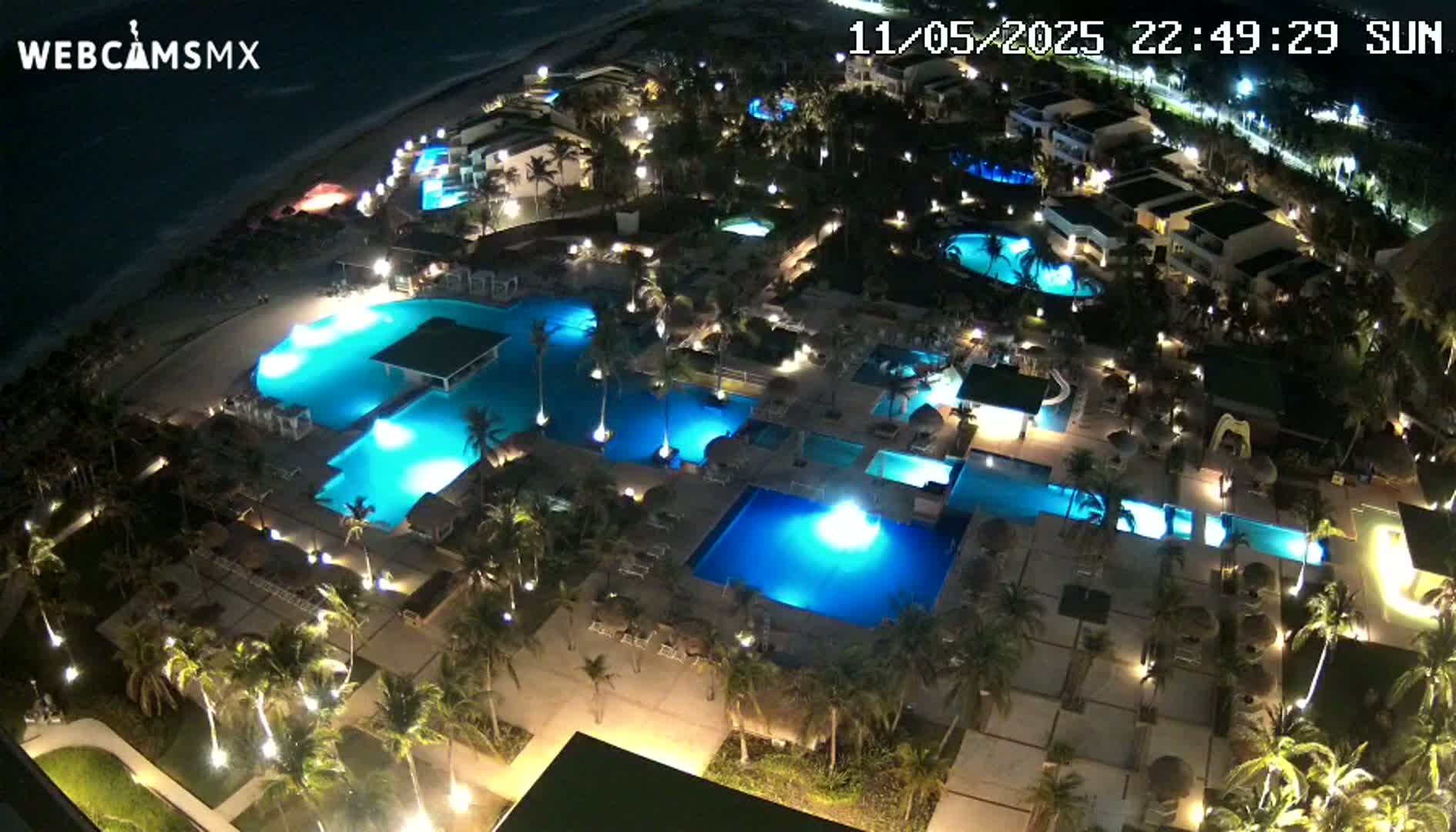 An aerial nighttime view shows a resort with numerous illuminated pools surrounded by palm trees and buildings near a beach under a dark sky.