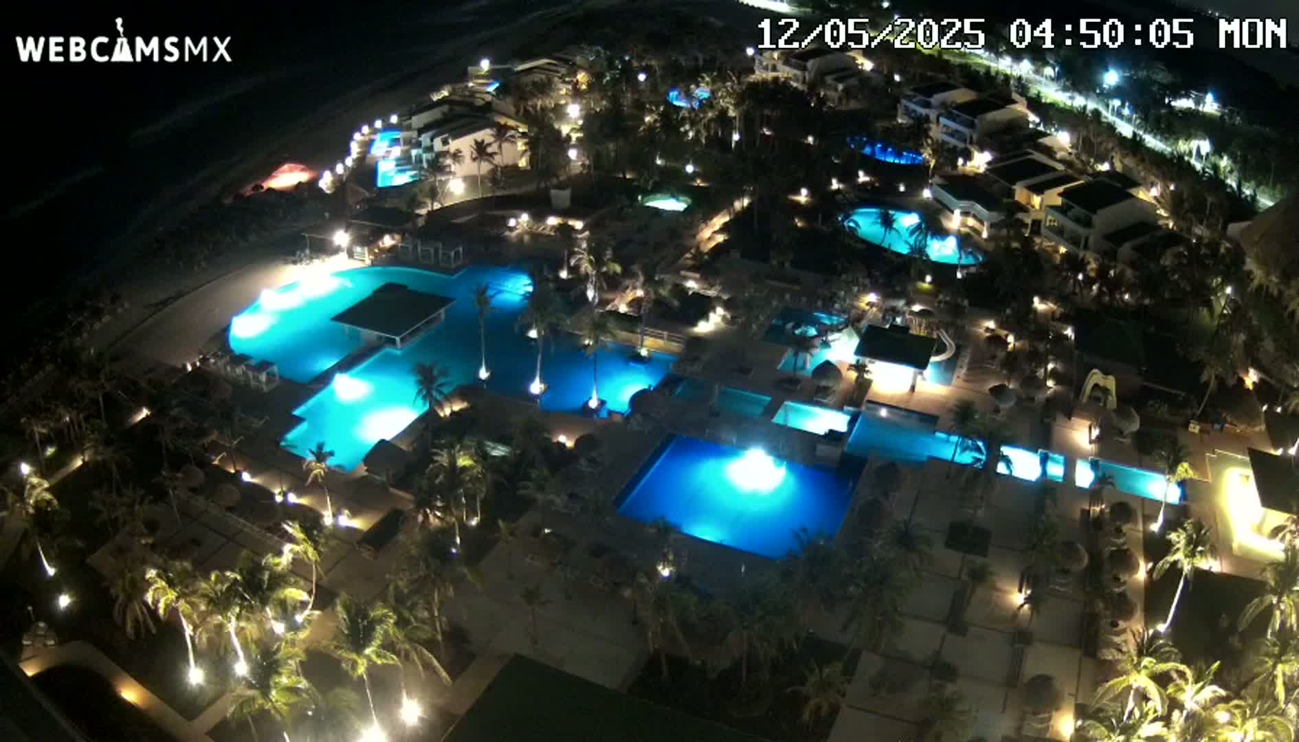 An aerial nighttime view of a resort with numerous illuminated swimming pools surrounded by palm trees and buildings.