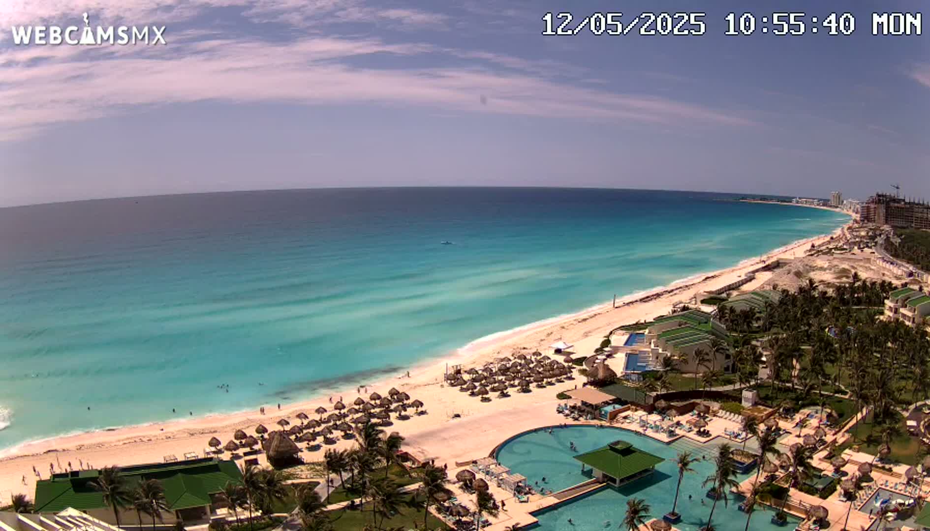 A mostly sunny day shows a long stretch of turquoise ocean meeting a white sand beach lined with beach umbrellas and palm trees, next to a resort with multiple pools.