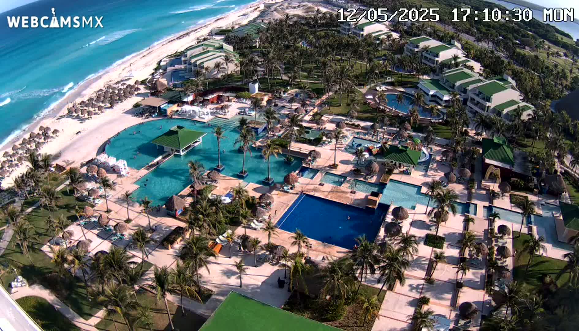 An aerial view shows a resort complex with numerous swimming pools, palm trees, and buildings situated next to a white sand beach and a turquoise ocean under sunny skies.