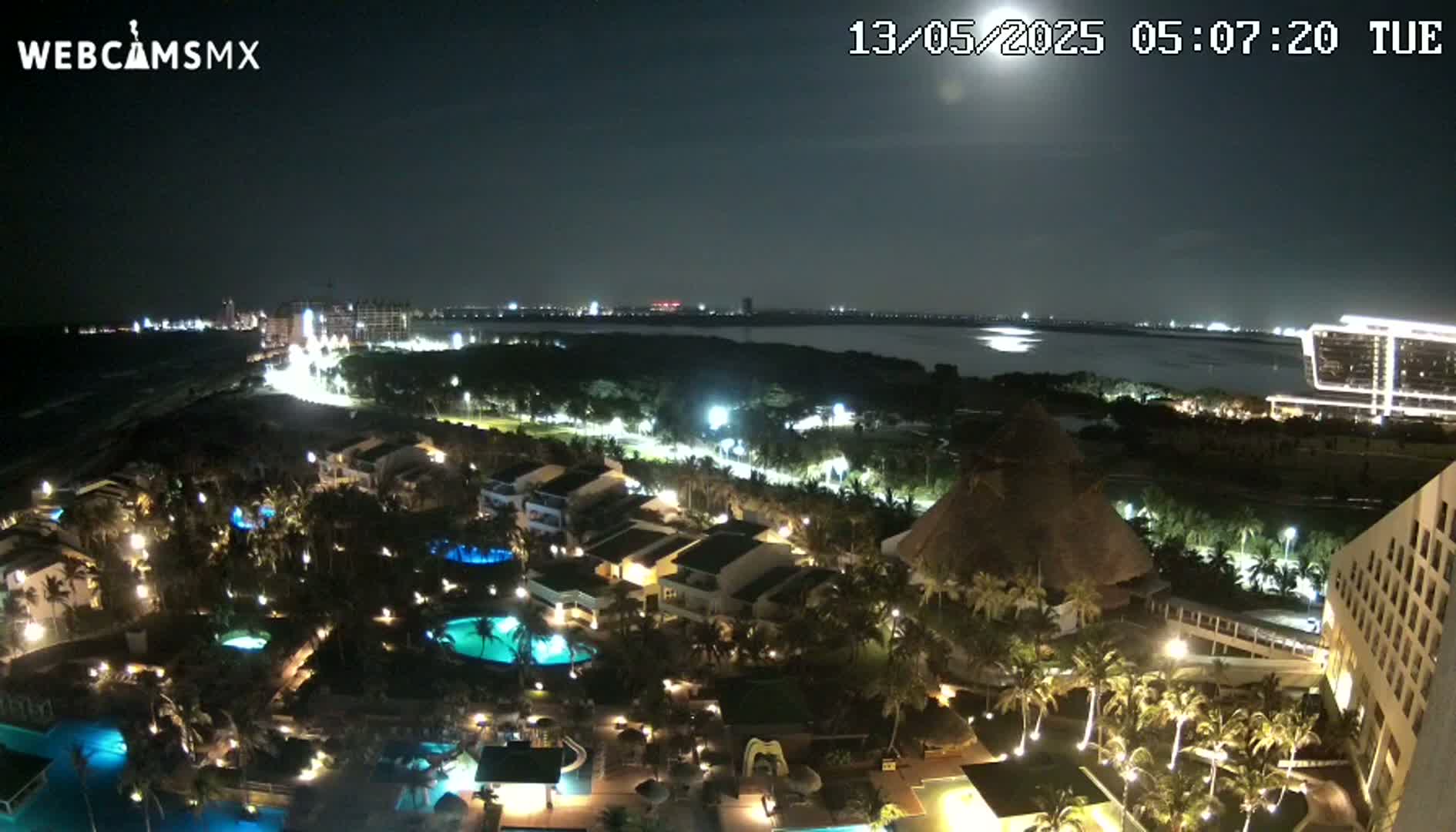 A nighttime aerial view of a resort complex with illuminated buildings, pools, and palm trees, overlooking a calm body of water under a bright moon.