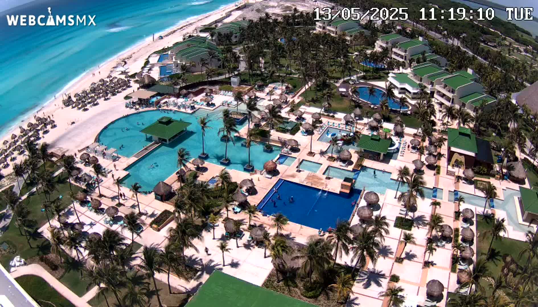 An aerial view shows a resort with numerous pools, palm trees, and a beach with turquoise water under sunny conditions.