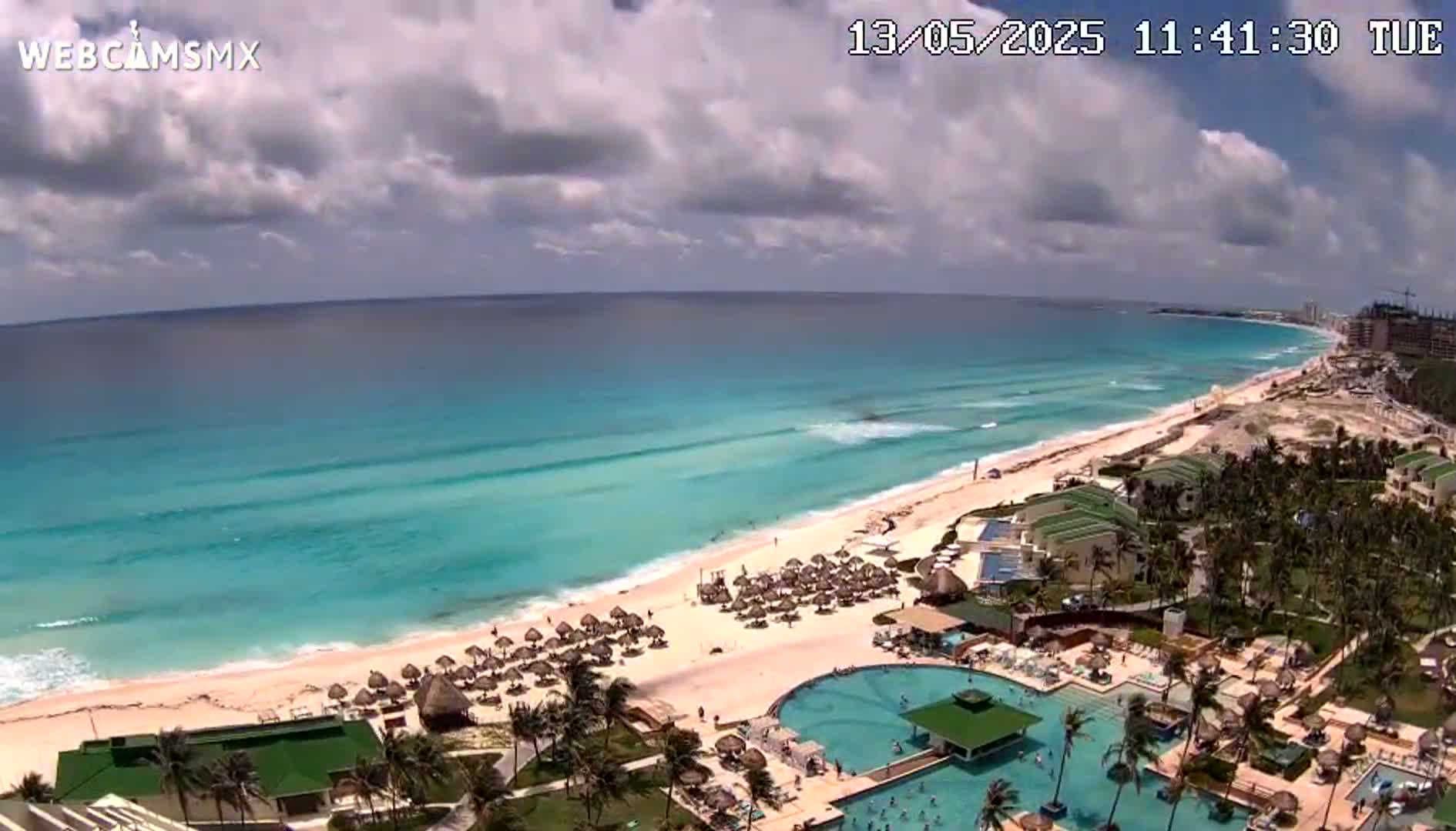An aerial view shows a partly cloudy day overlooking a long, sandy beach with turquoise water, resorts, and swimming pools.