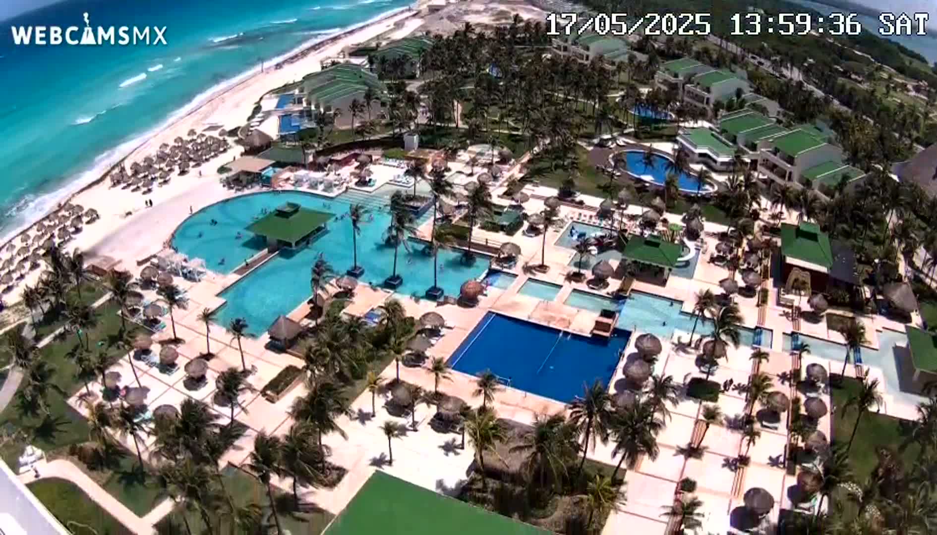 An aerial view shows a resort with numerous pools and palm trees surrounding a beach with turquoise water under sunny skies.