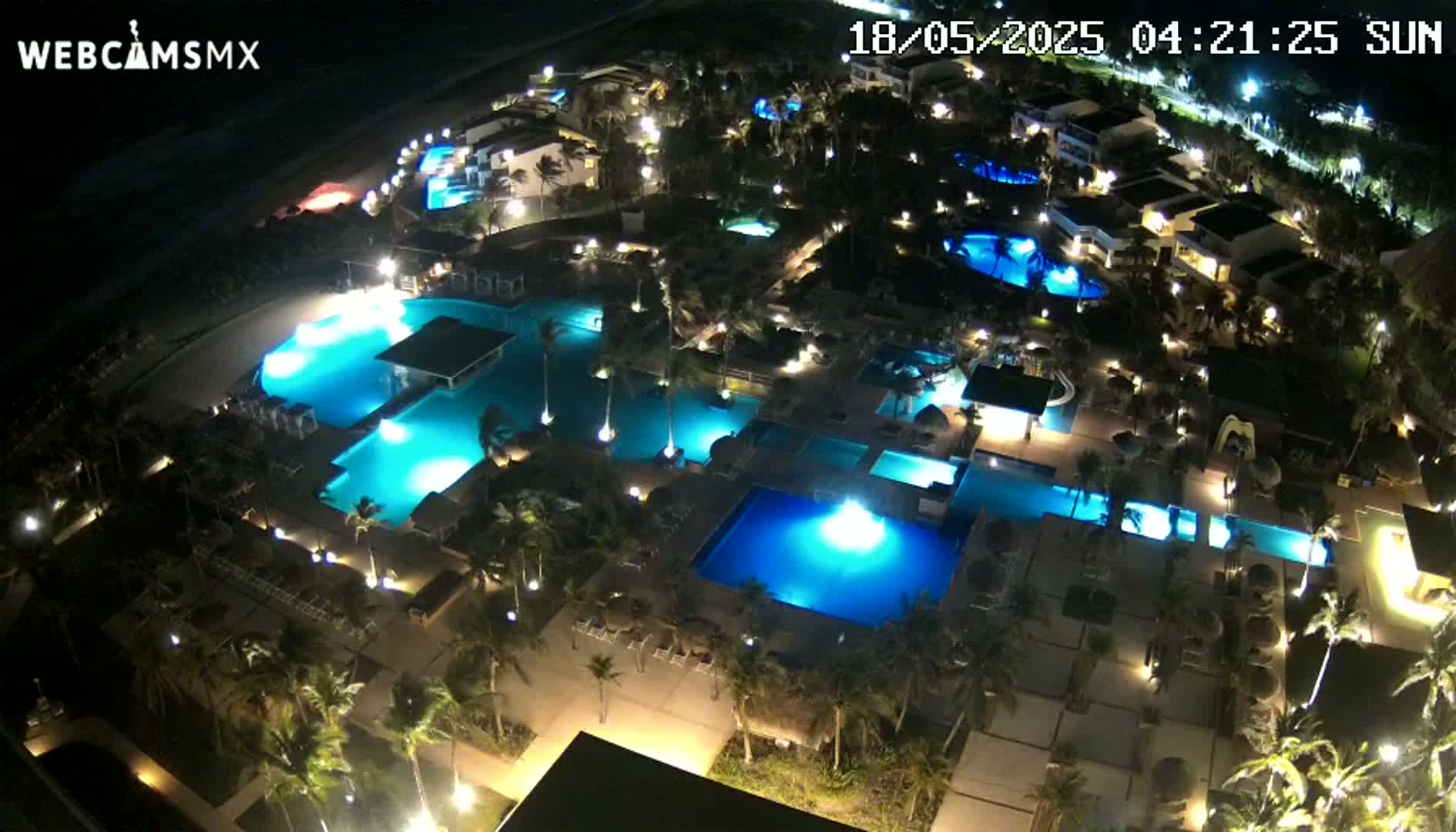 An aerial nighttime view of a resort complex with numerous illuminated swimming pools surrounded by palm trees and buildings.