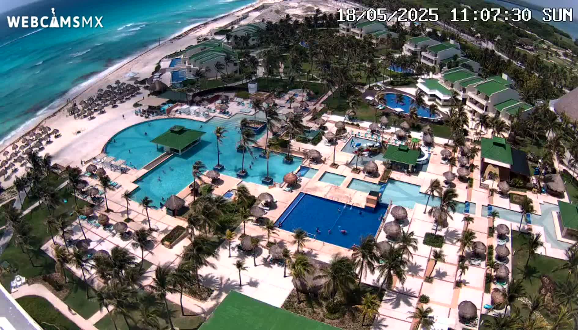 An aerial view shows a resort with numerous swimming pools, palm trees, and beach umbrellas surrounding low-rise buildings next to a sandy beach and turquoise ocean on a sunny day.