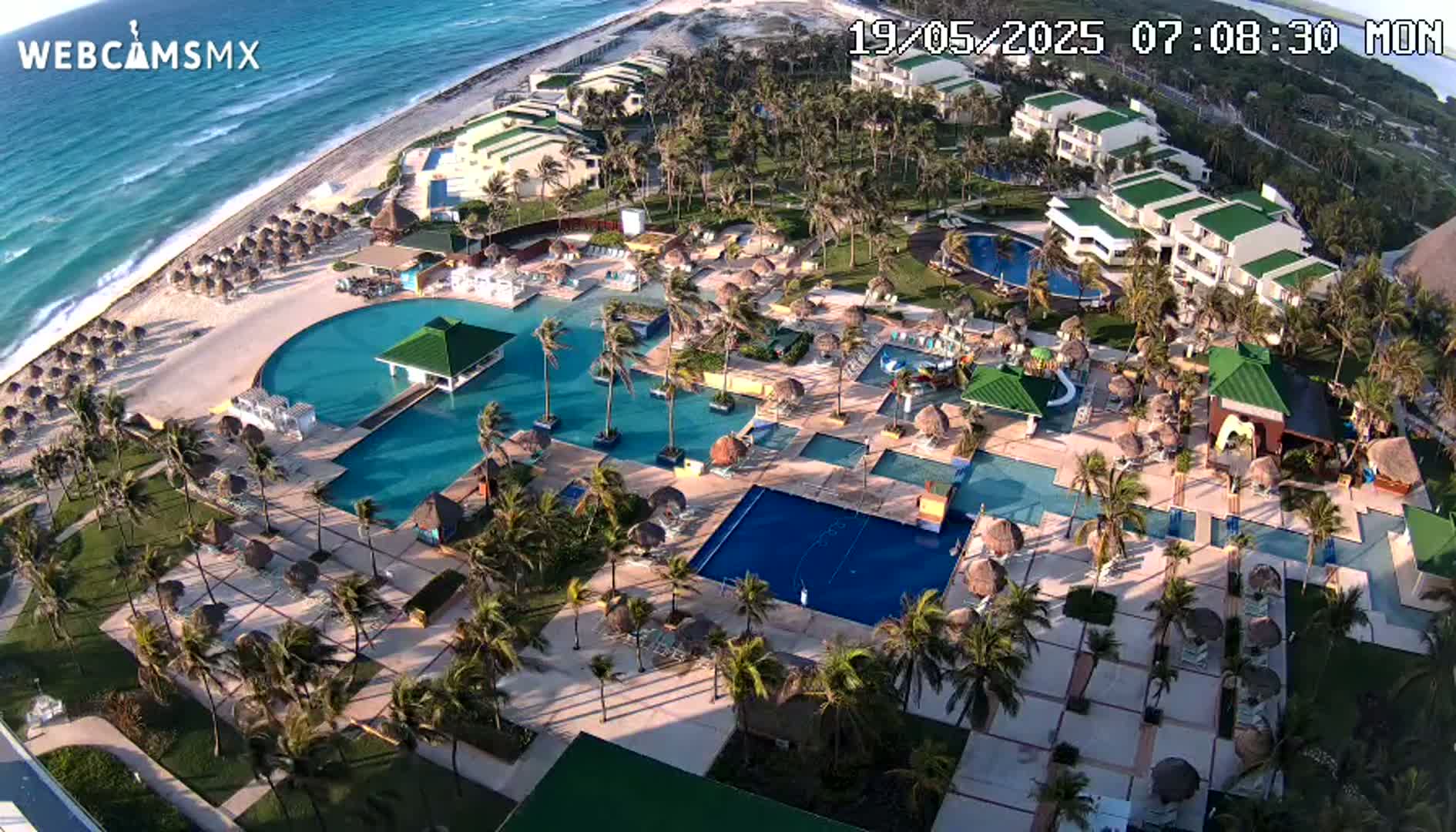 An aerial view shows a resort with numerous pools and palm trees situated next to a beach and ocean under sunny conditions.