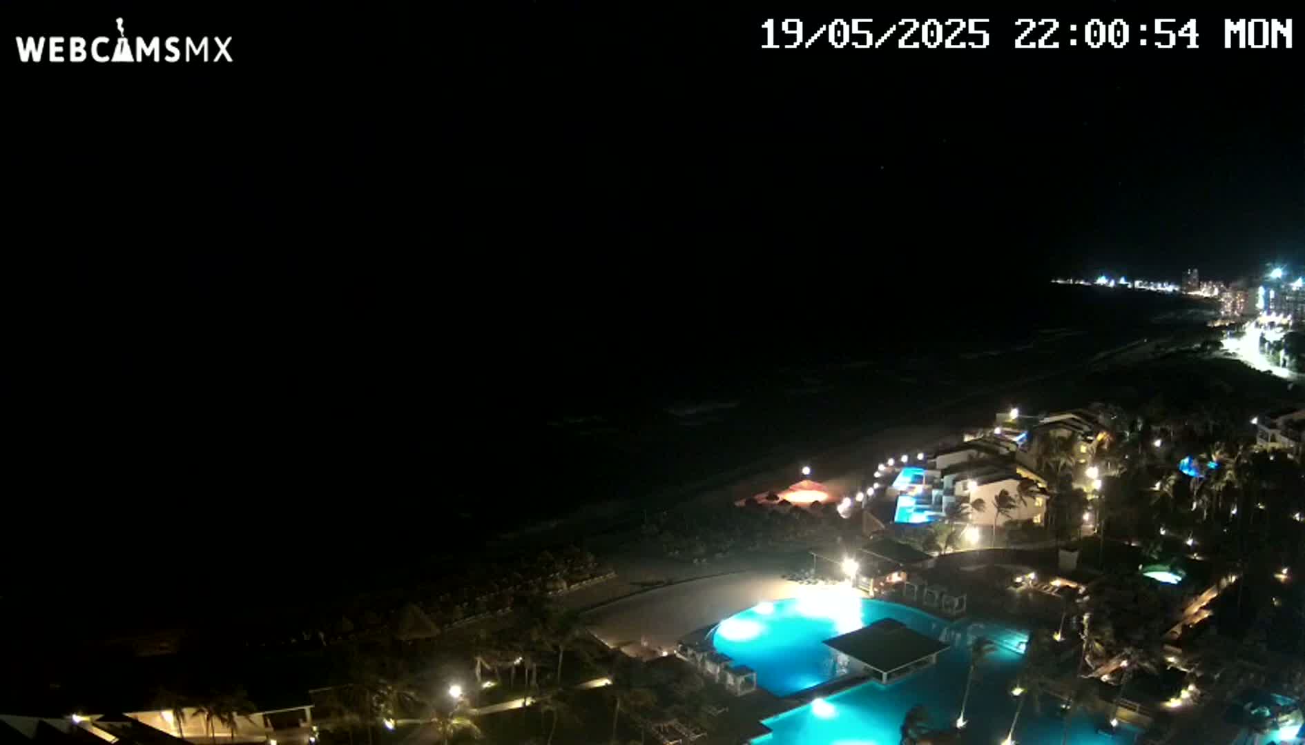 A nighttime, high-angle view shows a resort complex with illuminated pools and palm trees near a dark ocean under a clear, starless sky.
