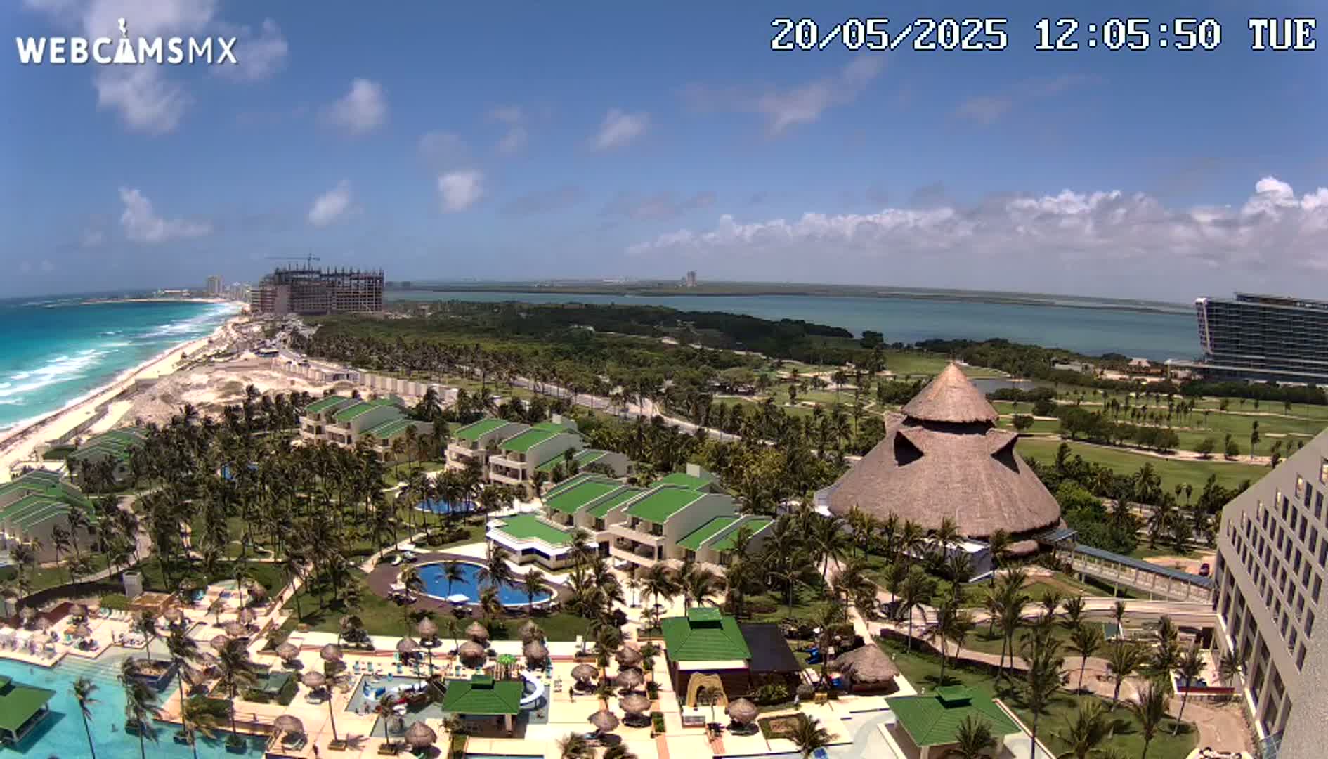A resort with multiple pools and buildings sits on a coast with a turquoise ocean under a partly cloudy sky.