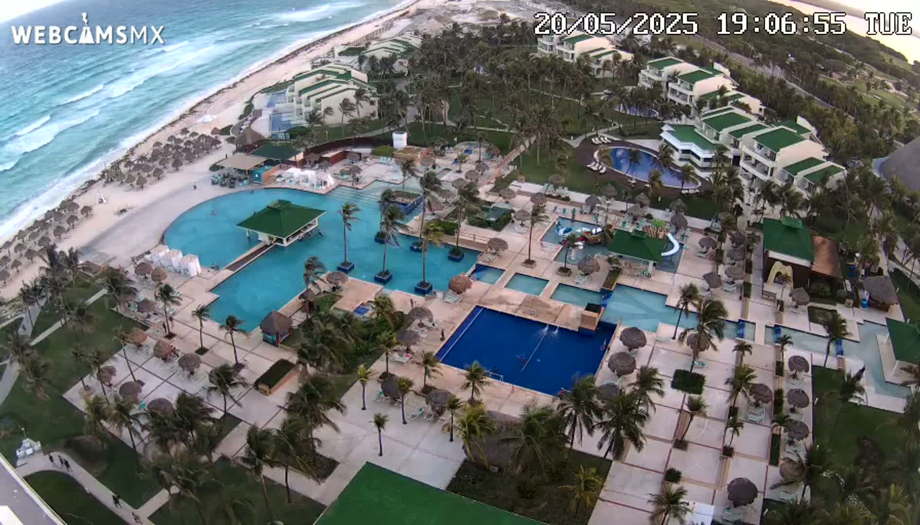 An aerial view shows a resort with multiple pools, surrounding buildings, and palm trees, next to a beach with ocean waves under a partly cloudy sky.