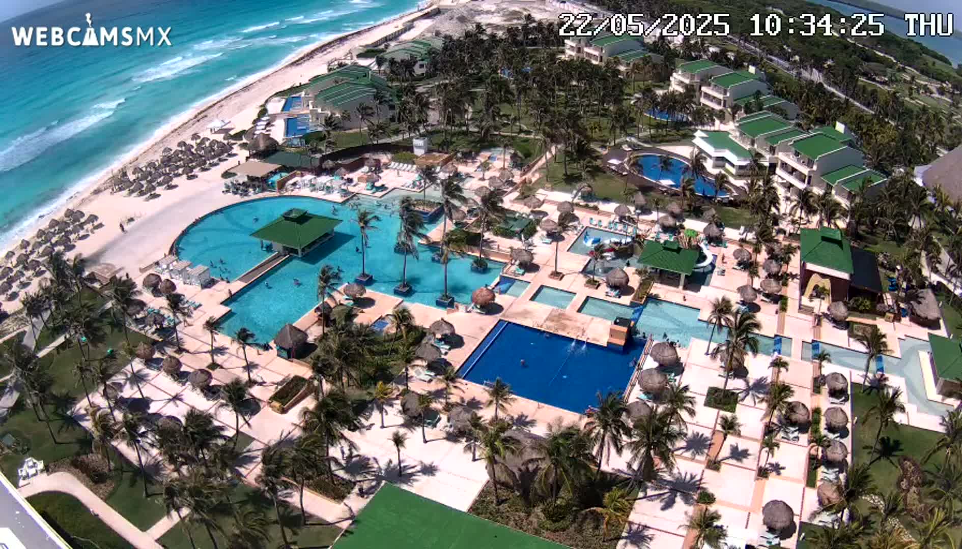 An aerial view shows a resort with multiple swimming pools, palm trees, and a beach with turquoise water under sunny skies.