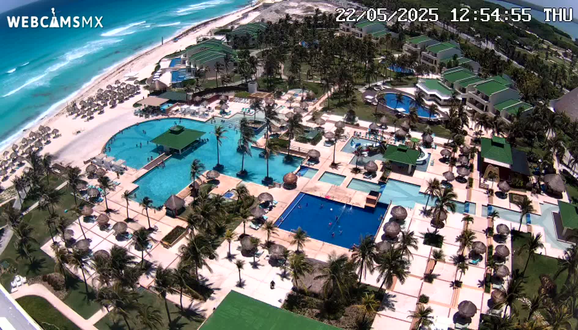 An aerial view shows a resort with numerous pools and palm trees next to a beach with turquoise water under a sunny sky.