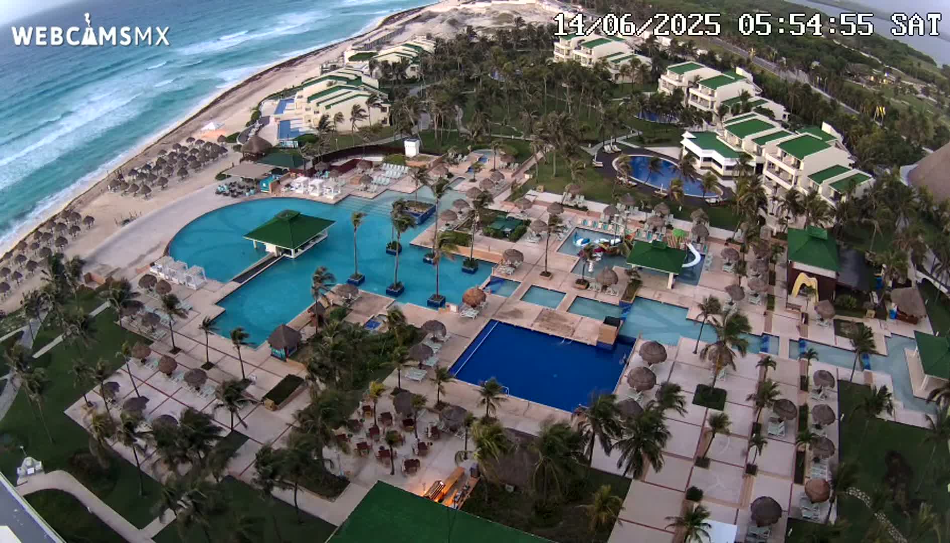 An aerial view shows a resort with multiple pools, beachside buildings, and palm trees, all under windy conditions near a wavy ocean.