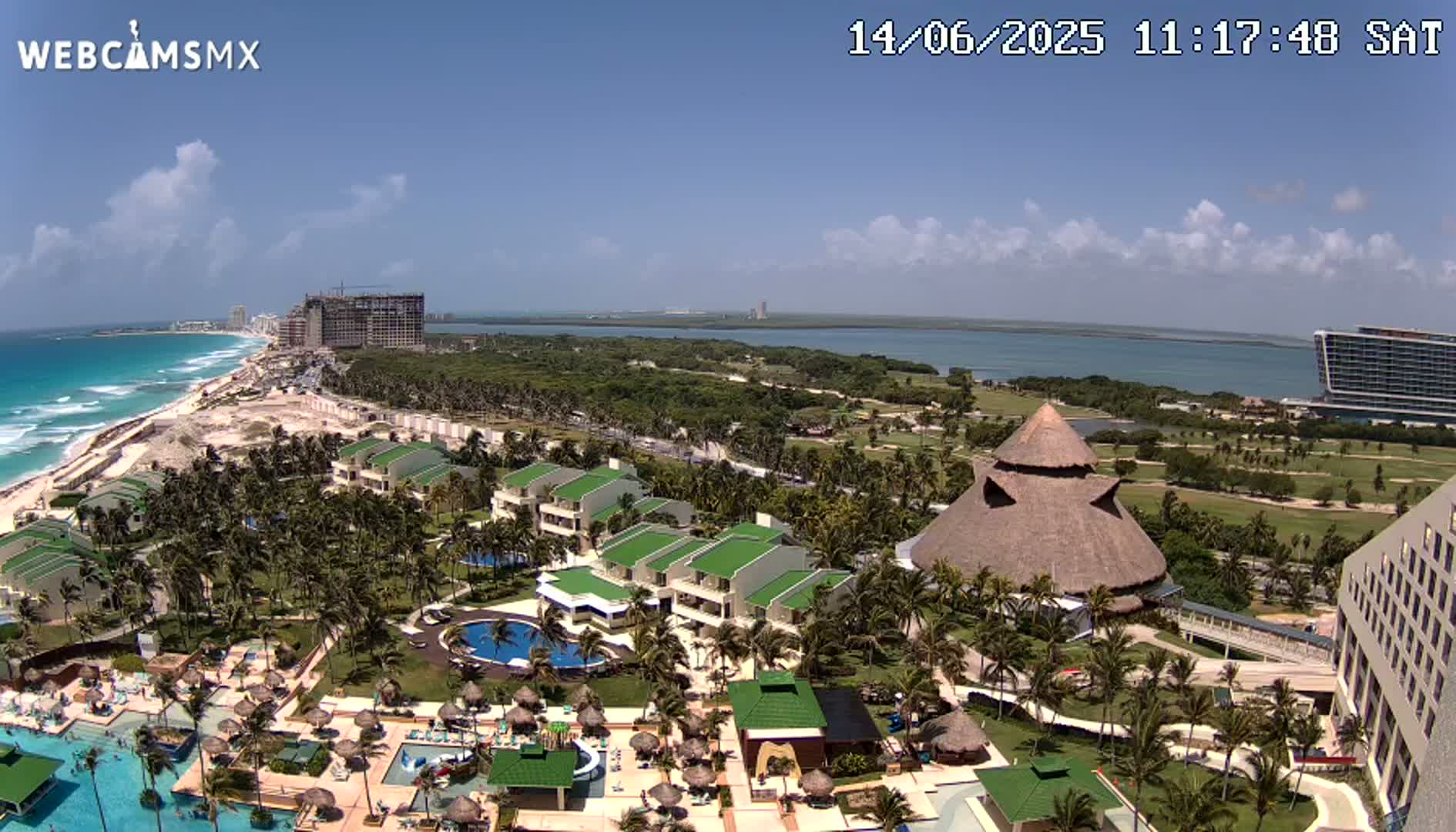 An aerial view shows a resort with pools and palm trees next to a beach on a sunny day, with a body of water and more hotels in the distance.