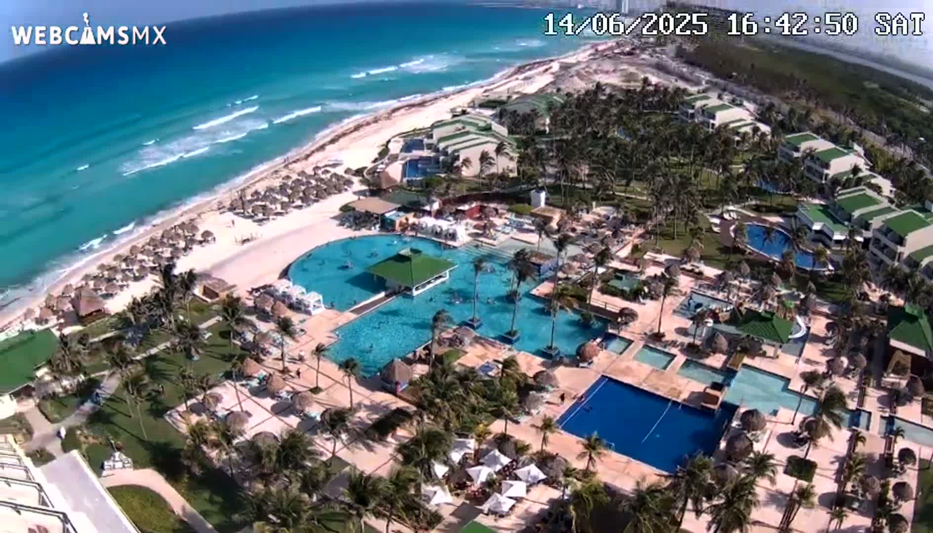 An aerial view shows a resort with pools and palm trees next to a beach on a sunny day, with a body of water and more hotels in the distance.