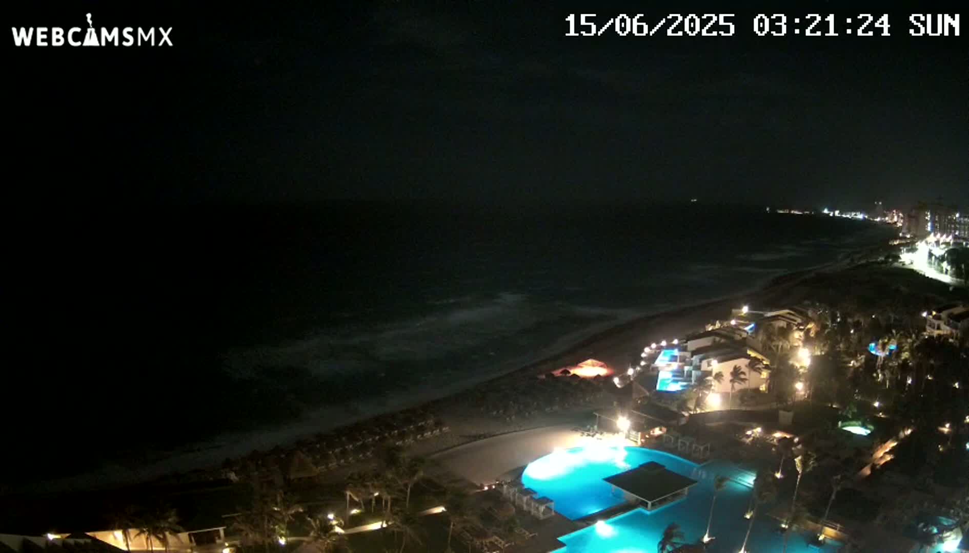 A nighttime, high-angle view shows a resort's illuminated swimming pools and buildings next to a dark ocean with small waves under a mostly clear night sky.