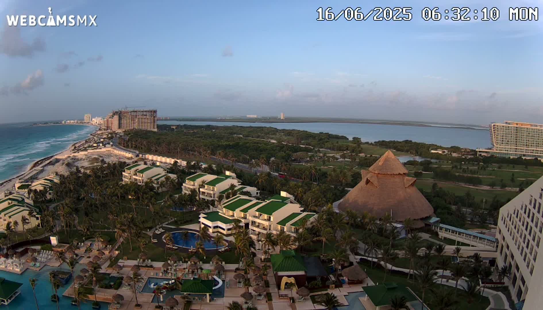 An aerial view of a tropical resort area shows a beach, ocean, several buildings, swimming pools, lush vegetation, and a clear, sunny sky.