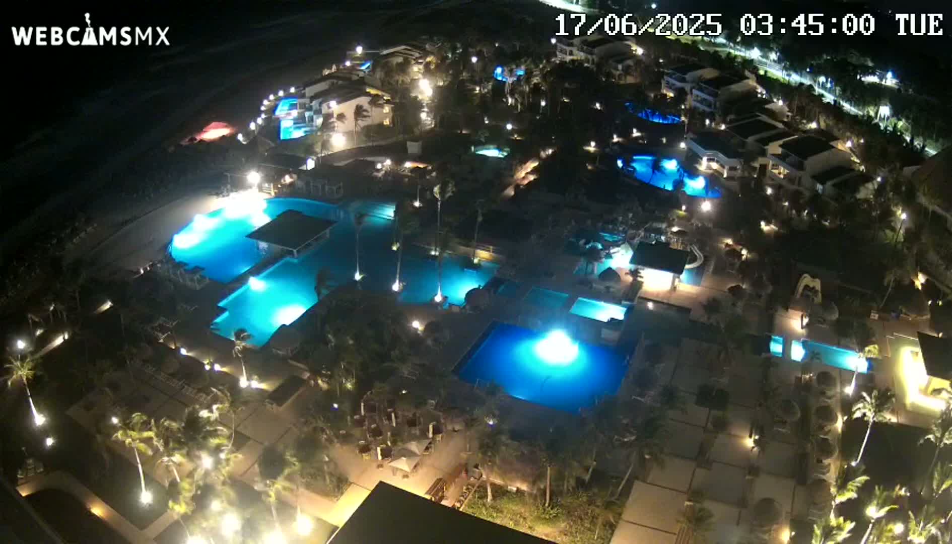 An aerial nighttime view of a resort with numerous illuminated swimming pools surrounded by palm trees and buildings.