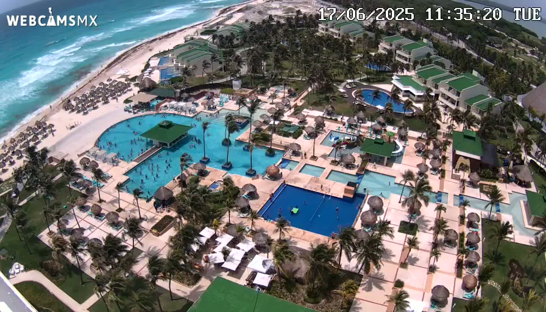 An aerial view shows a resort with numerous pools, surrounding landscaping, and a beach with turquoise water under a sunny sky.