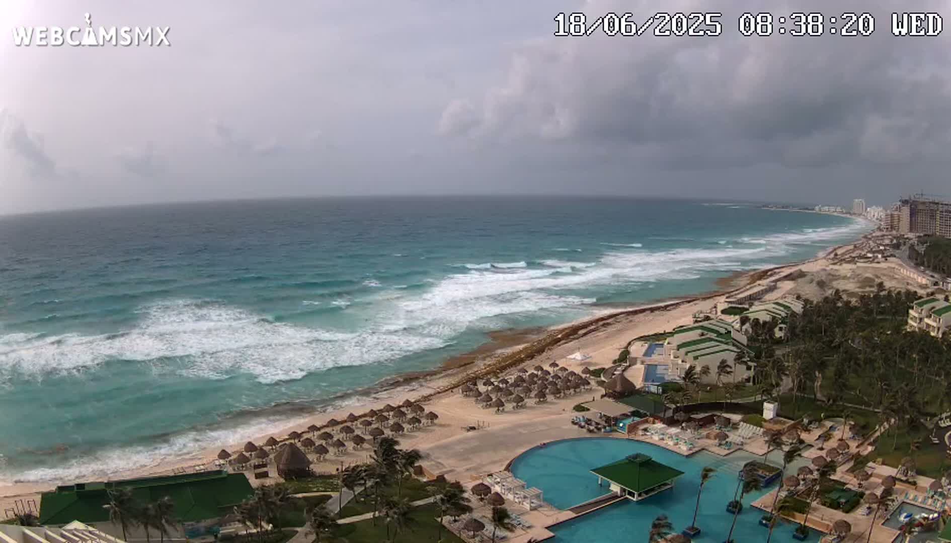 A resort with a large pool overlooks a beach with moderate waves under a cloudy sky.