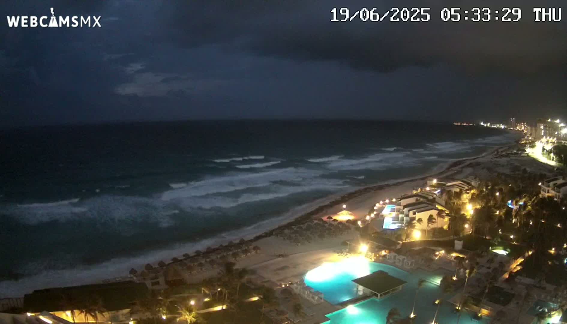 A nighttime view of a beach resort with a large pool illuminated under a dark, stormy sky, showing waves crashing on the shore.