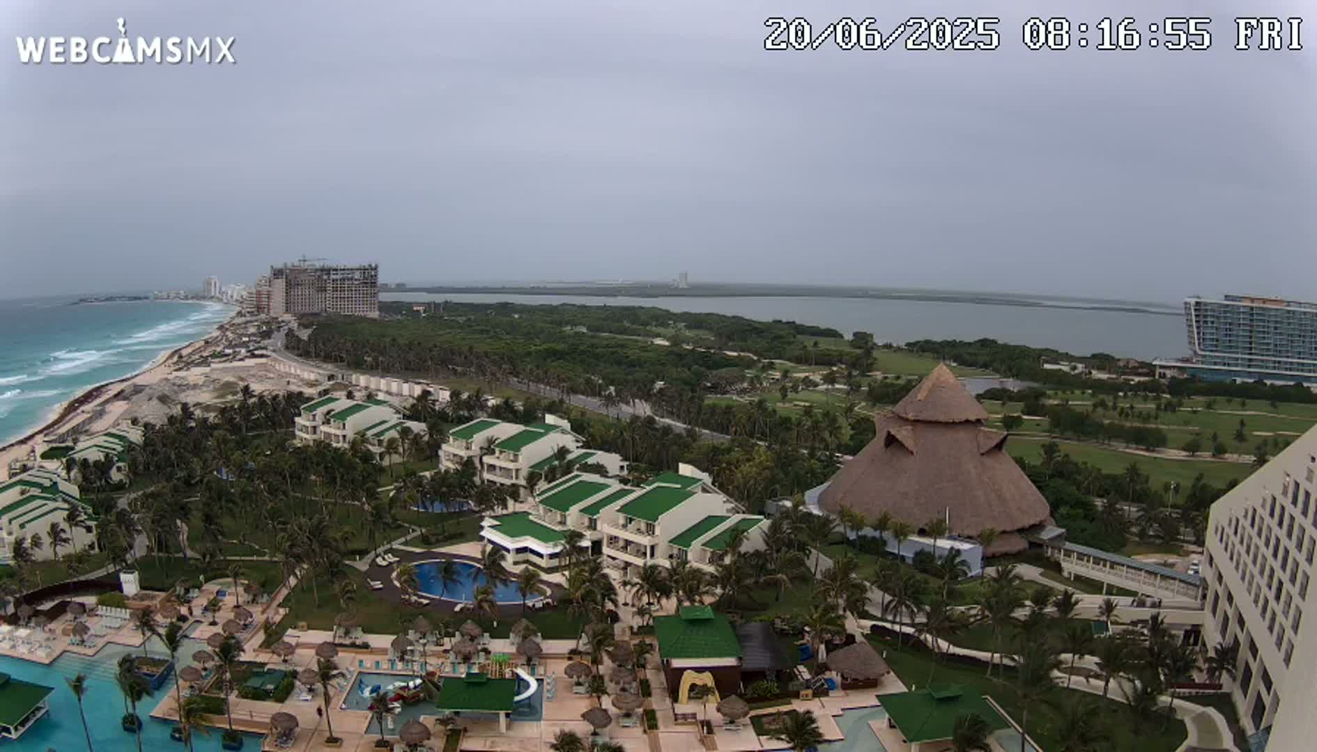 An aerial view shows a tropical resort with multiple buildings, pools, and palm trees, situated on a coast with a beach and ocean under an overcast sky.