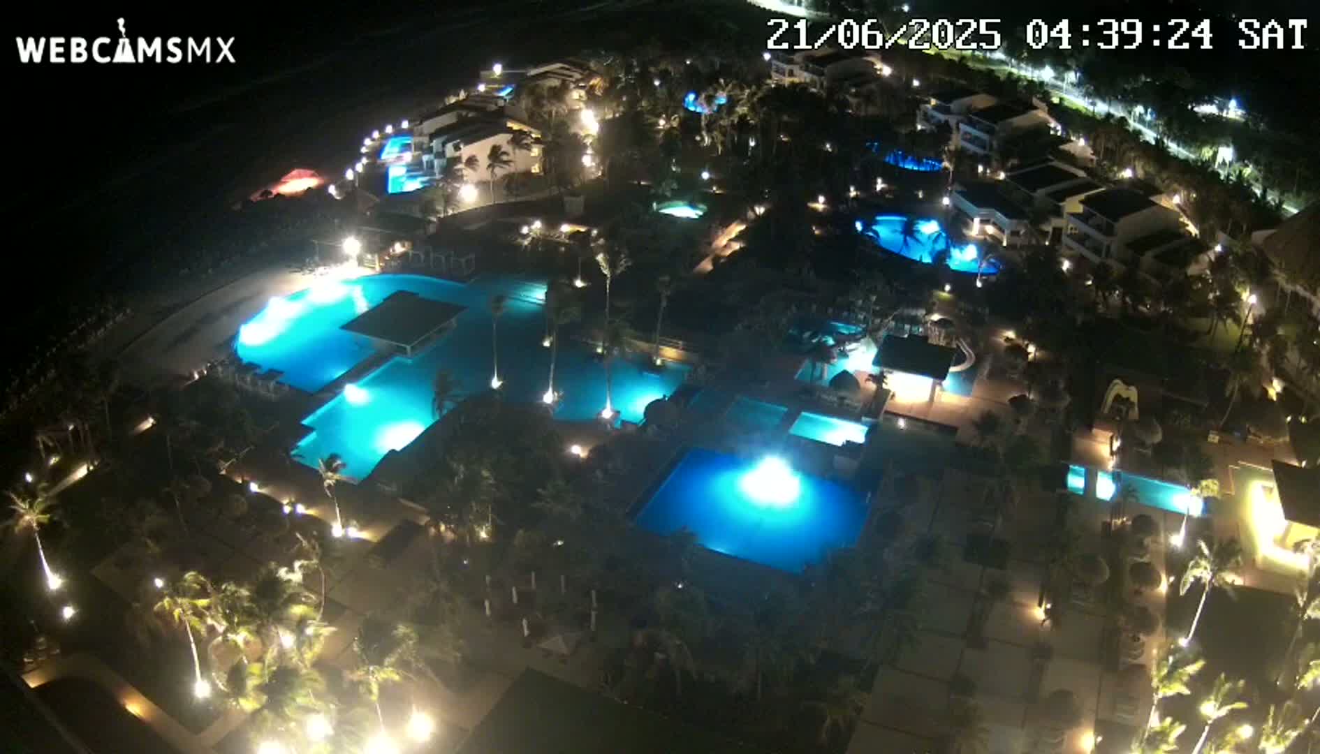An aerial nighttime view of a resort complex with numerous illuminated swimming pools surrounded by palm trees and buildings.
