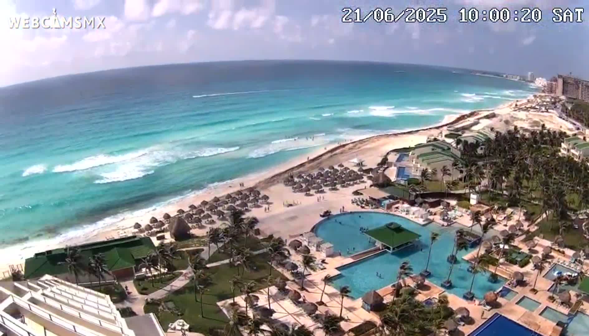 An aerial view shows a resort with multiple pools and beach umbrellas next to a turquoise ocean with moderate waves under a partly sunny sky.