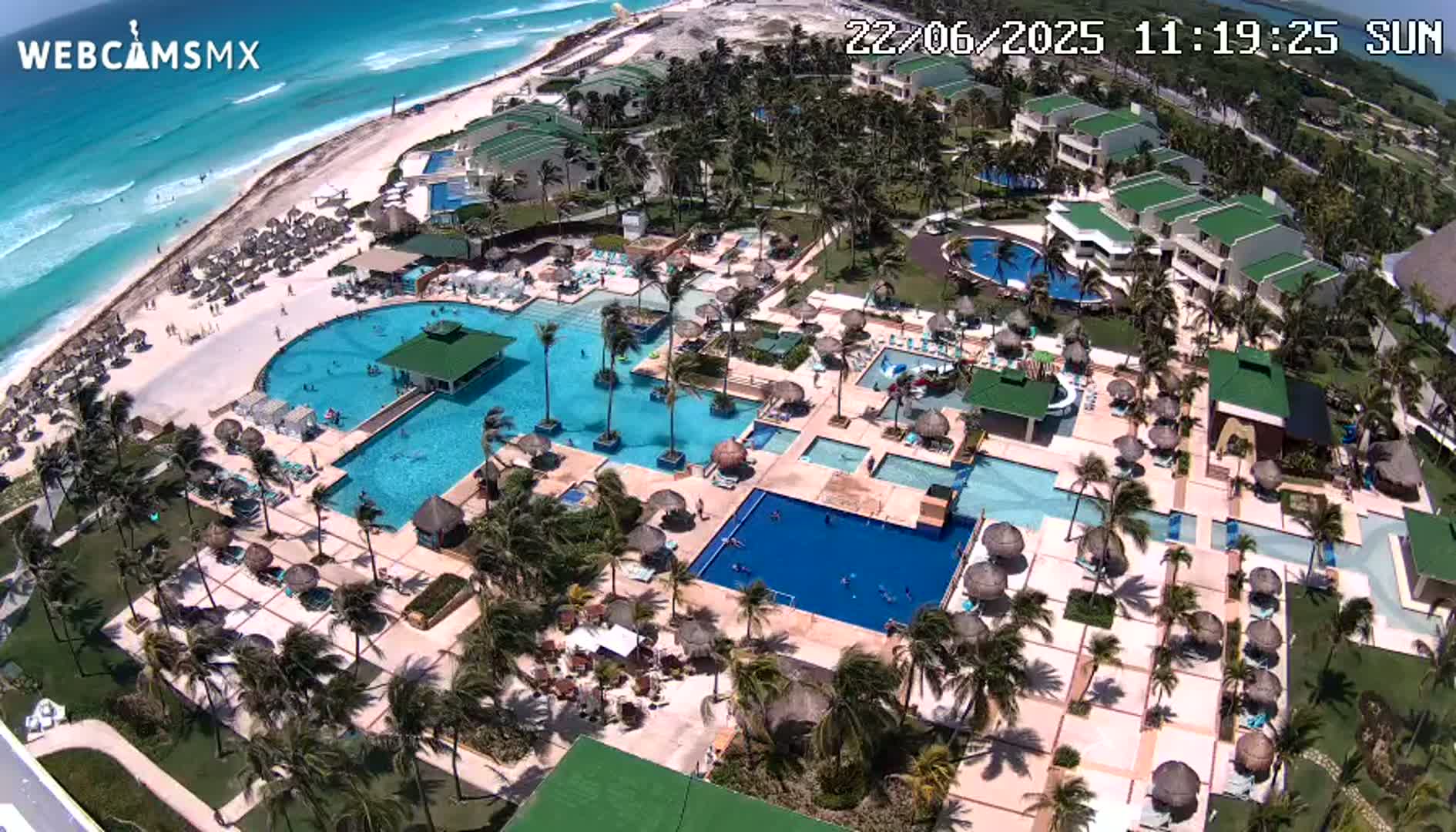 An aerial view shows a resort with multiple pools and buildings surrounded by palm trees, adjacent to a beach and turquoise ocean on a sunny day.