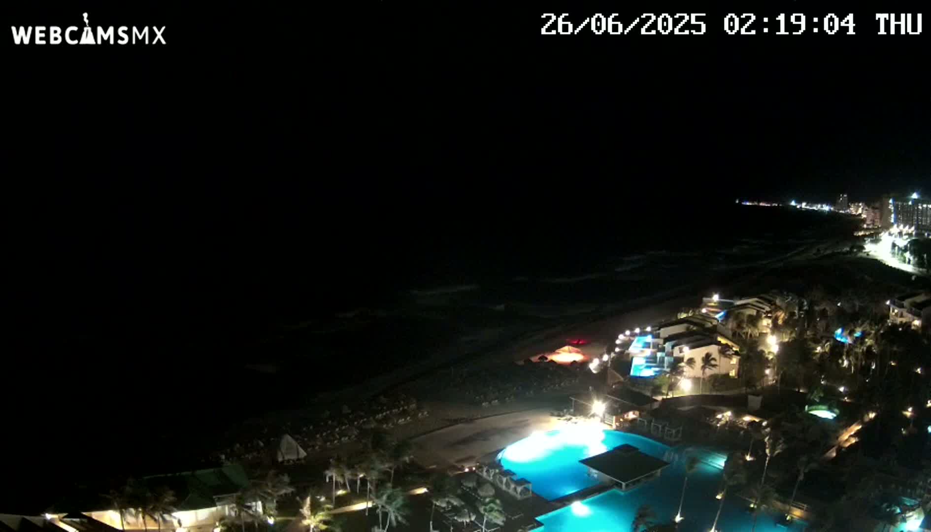 A nighttime, aerial view shows a resort complex with illuminated pools and buildings along a dark ocean shoreline under a clear sky.