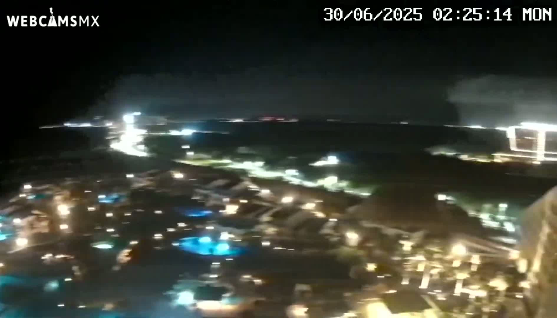 A nighttime aerial view shows a resort area with illuminated pools and buildings under a mostly cloudy sky.